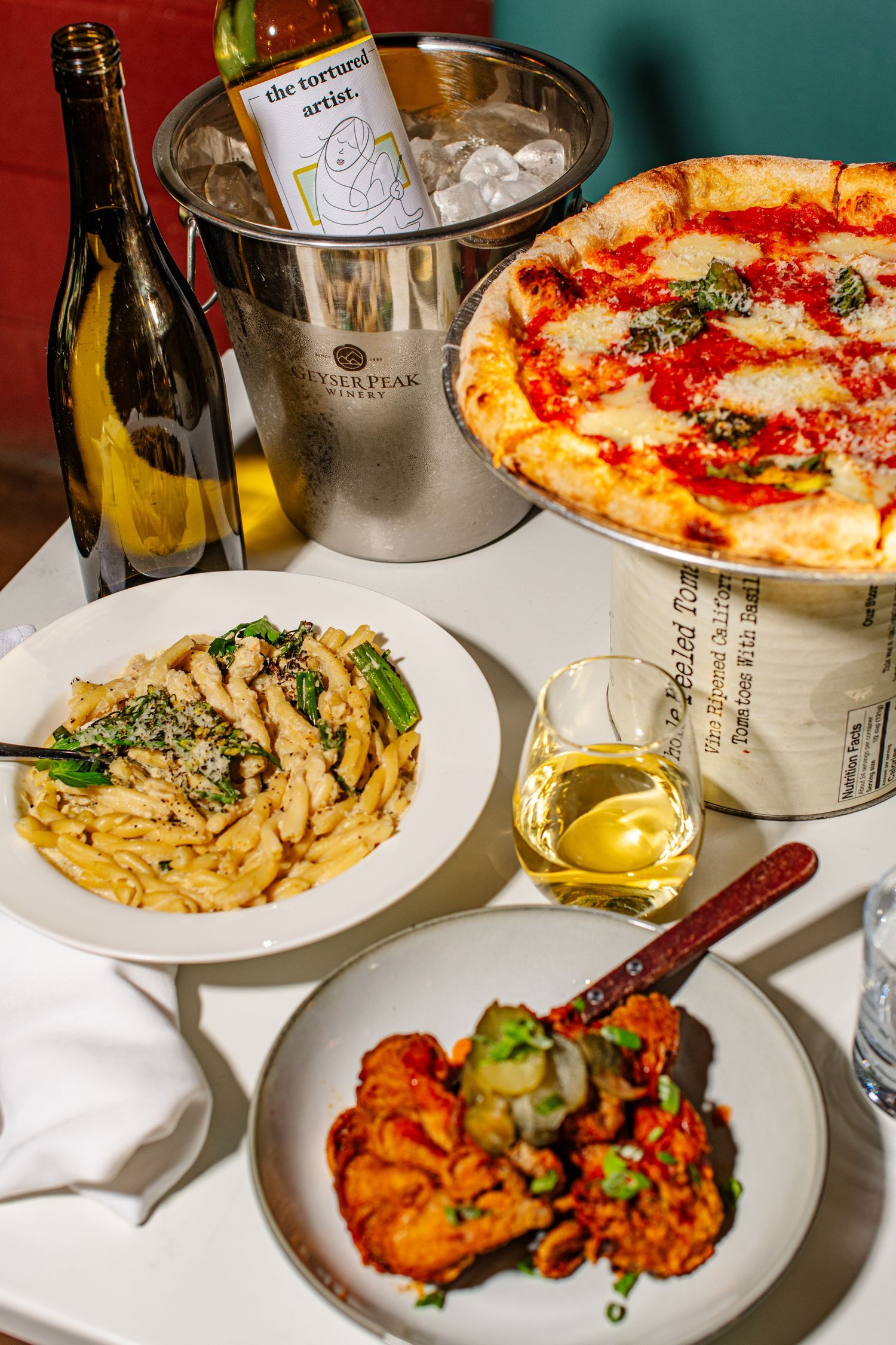 A table spread with Italian dishes including pasta, pizza, and grilled meats alongside wine glasses and white plates.