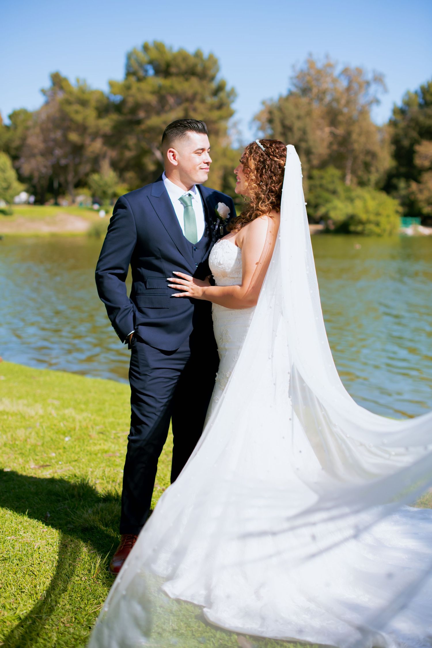 A wedding couple poses together by a scenic Legg Lake on a sunny day, with the bride's veil flowing in the breeze.