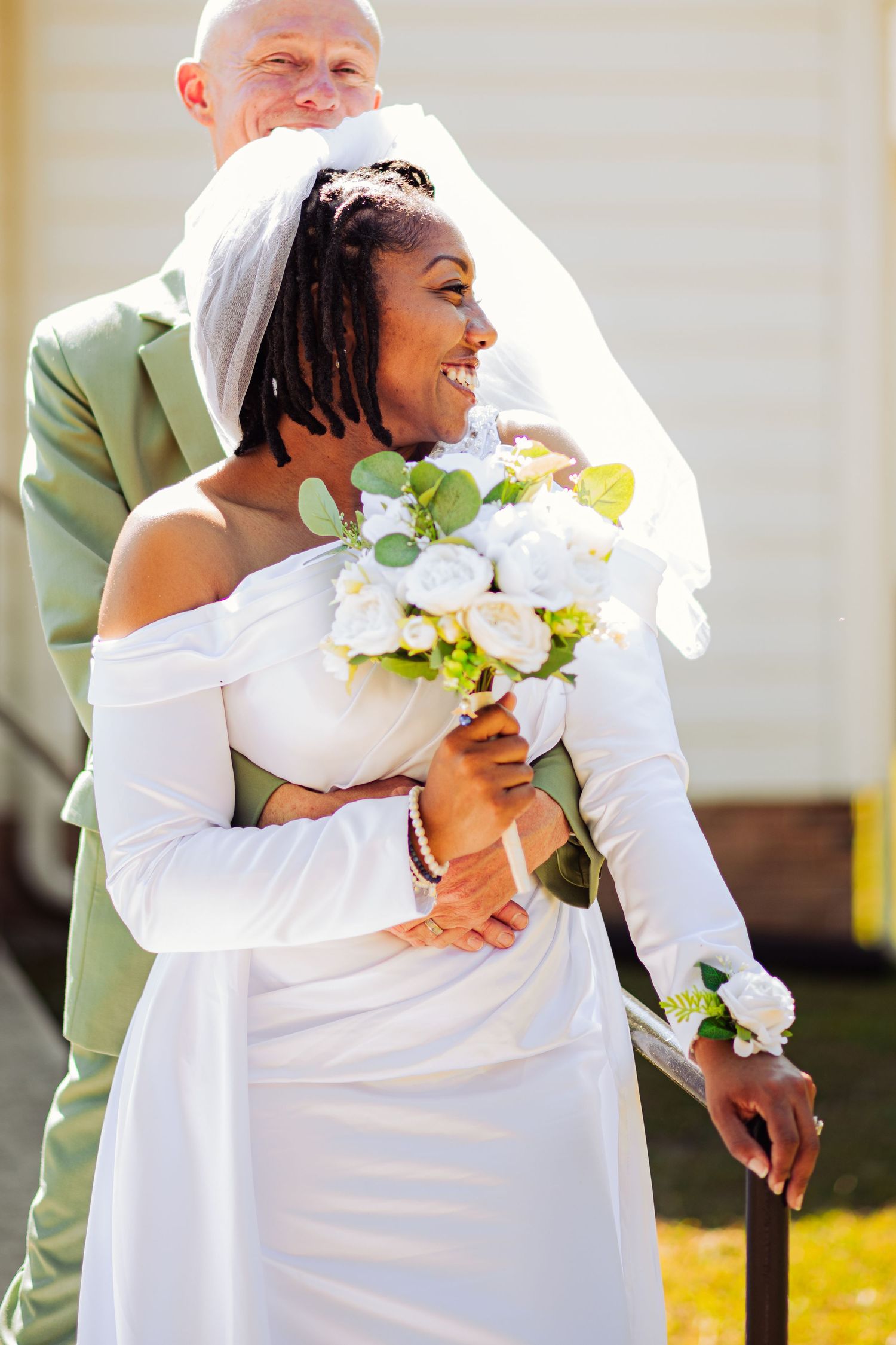 Wedding couple embraces outdoors while wearing white dress and sage green attire.