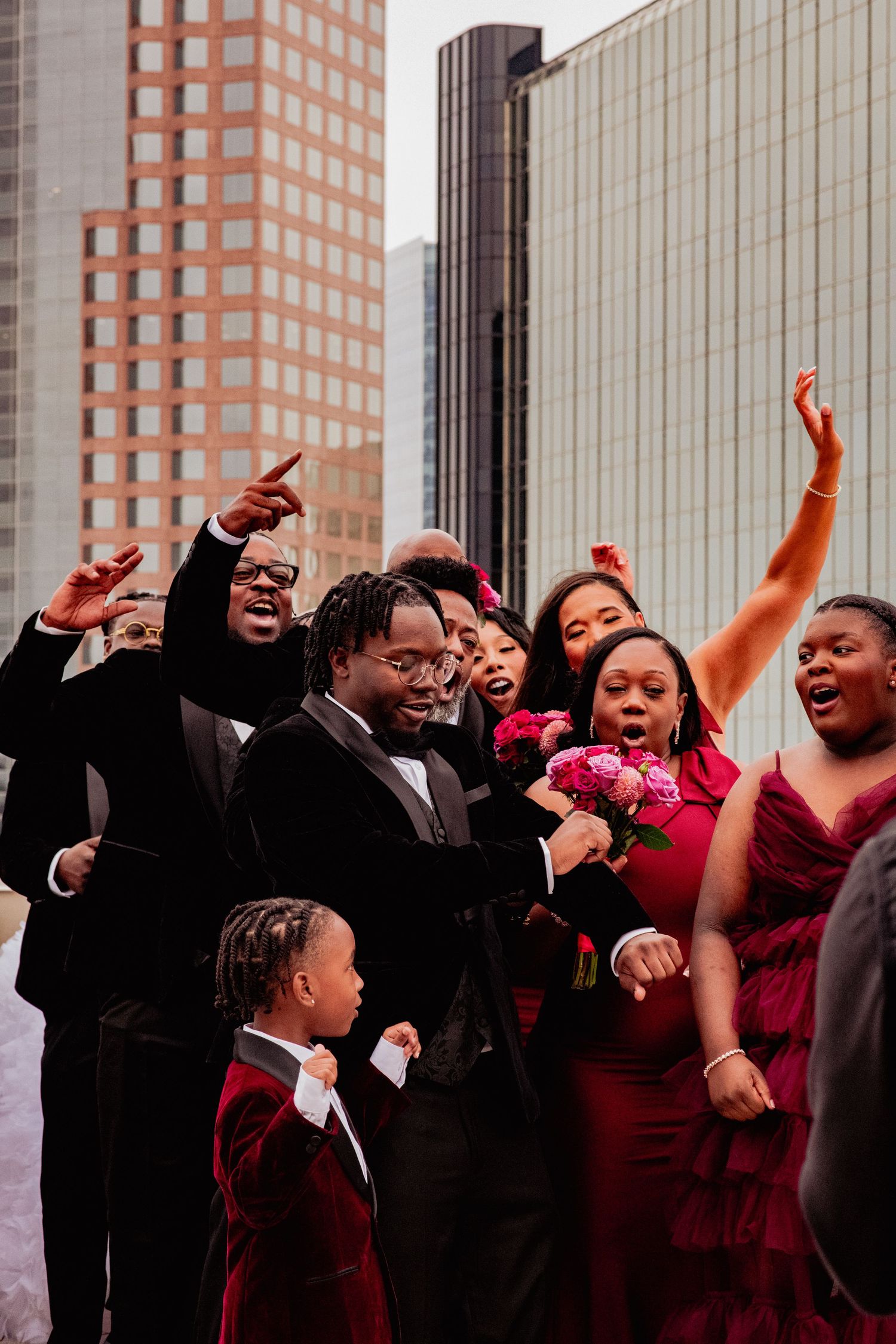 Wedding party in burgundy and black attire celebrating joyfully against urban architecture backdrop.