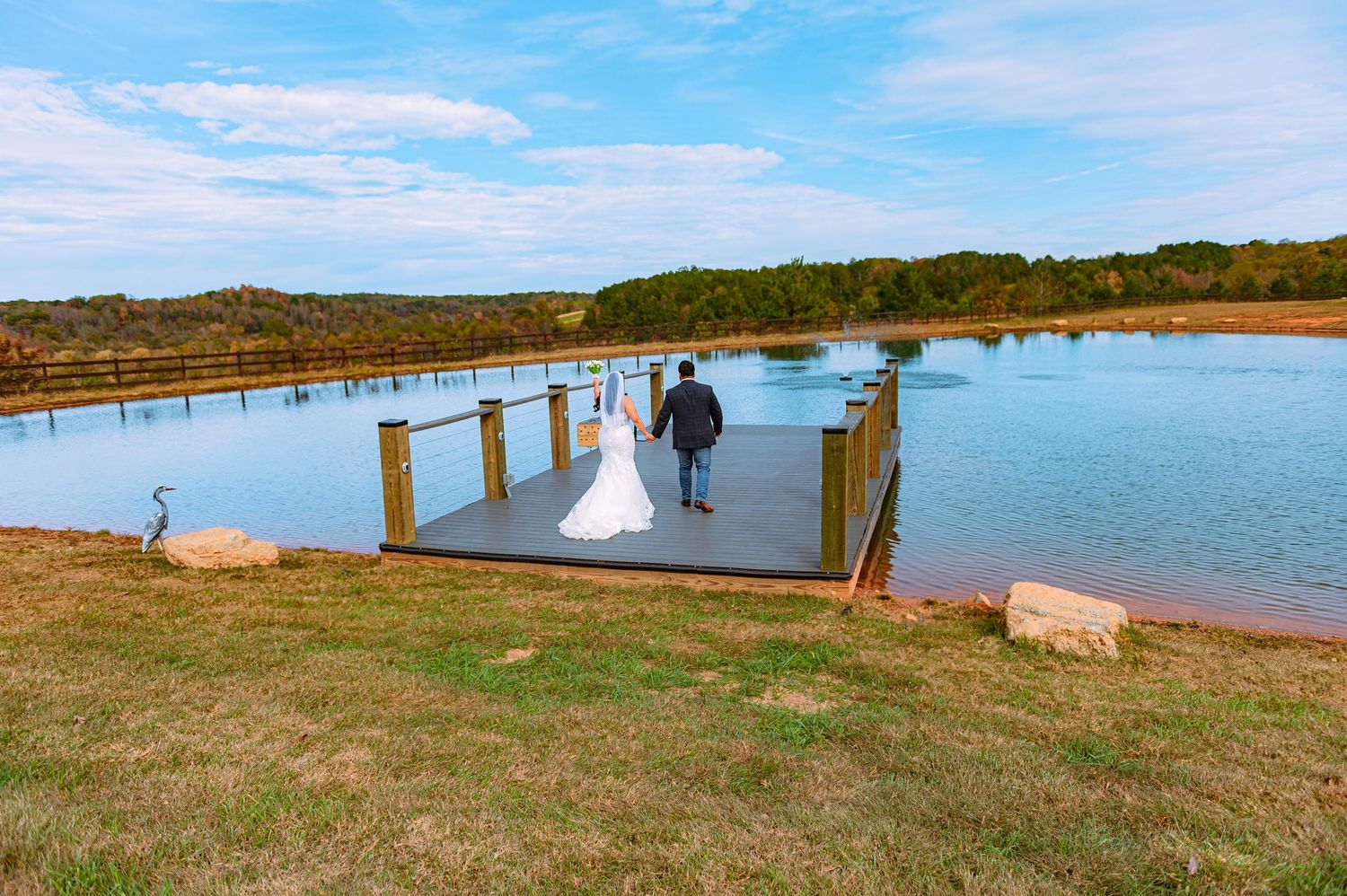 Panoramic view of a wooden boardwalk along a coastal shoreline with blue water and grass-covered hills in the background.