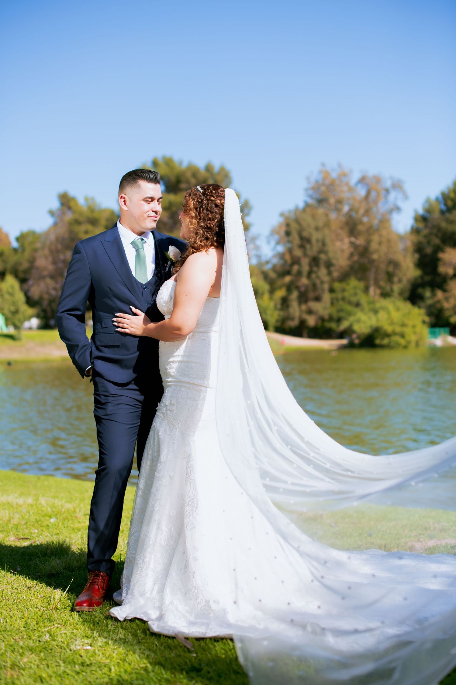 Lakeside wedding portraits with dramatic flowing veil against blue sky and water.