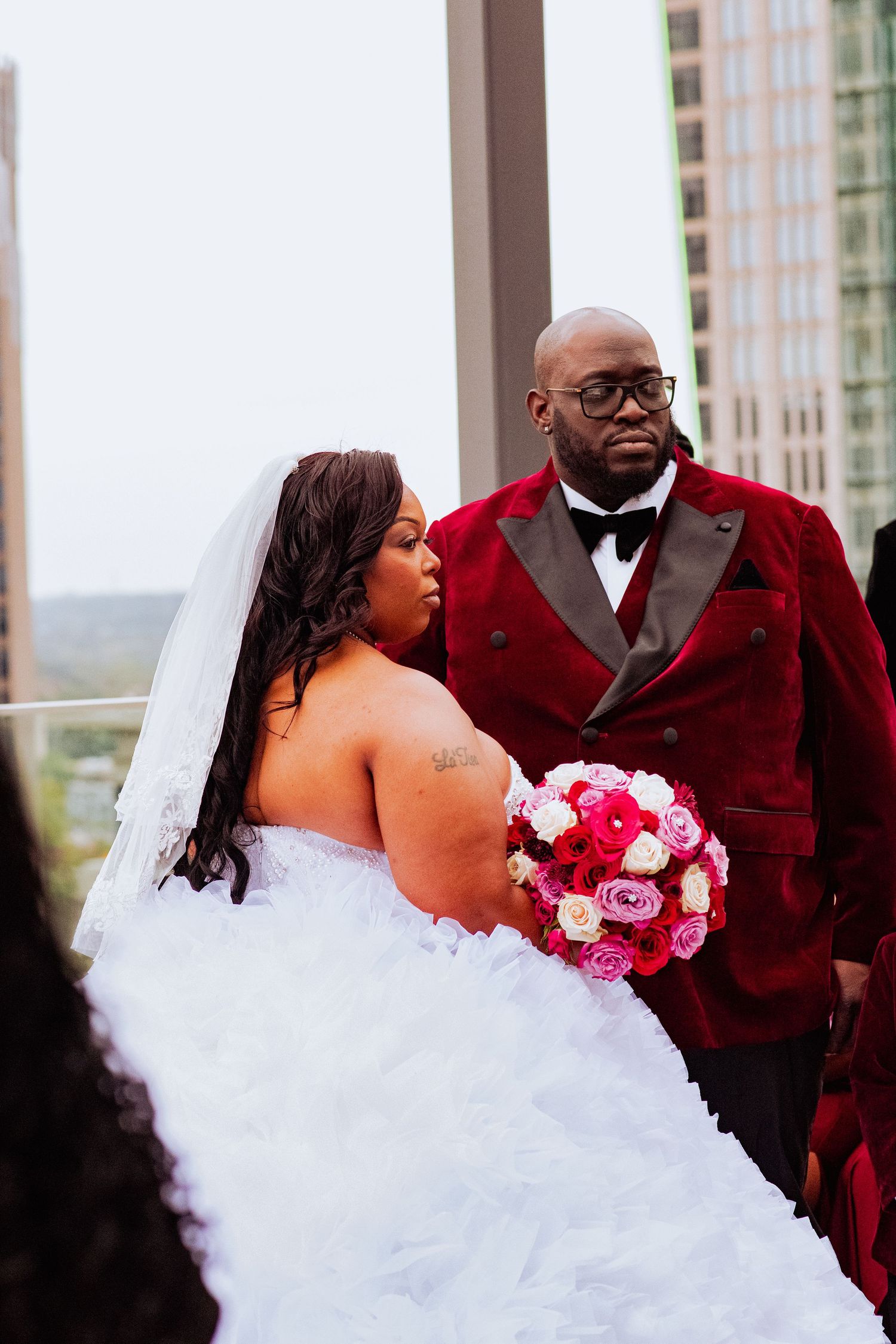 Wedding couple shares candid moments in white gown and burgundy suit against city skyline.