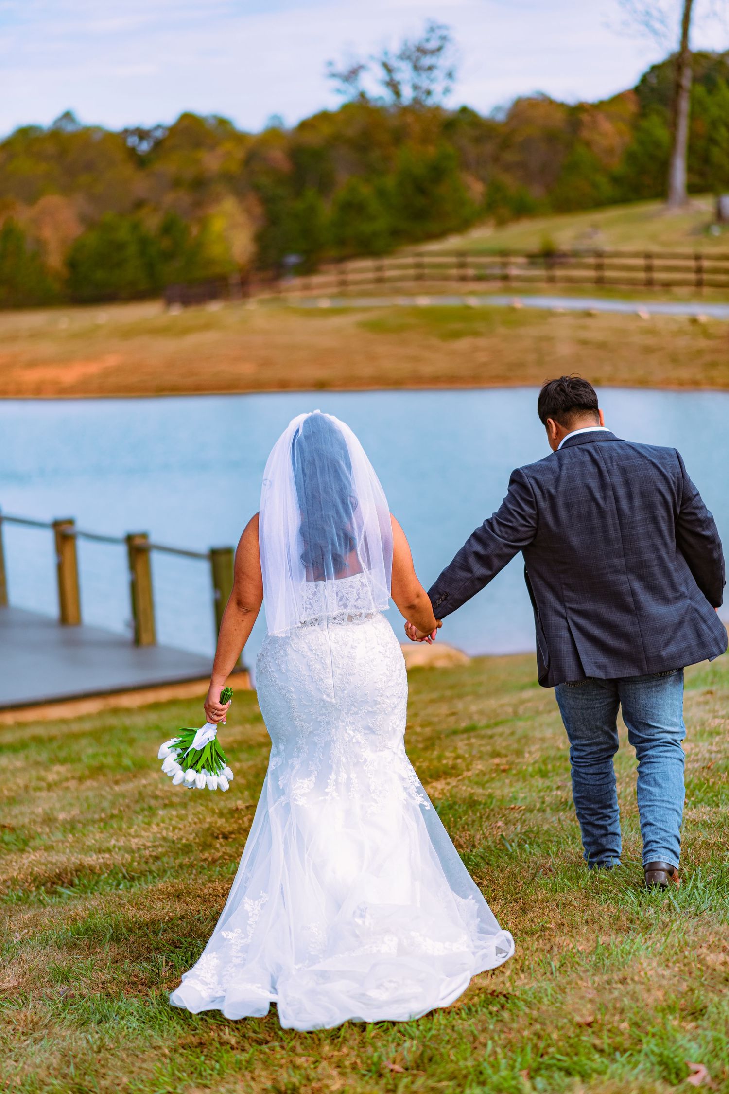 A couple in wedding attire walking together on a grassy hill overlooking a serene lake on their special day.