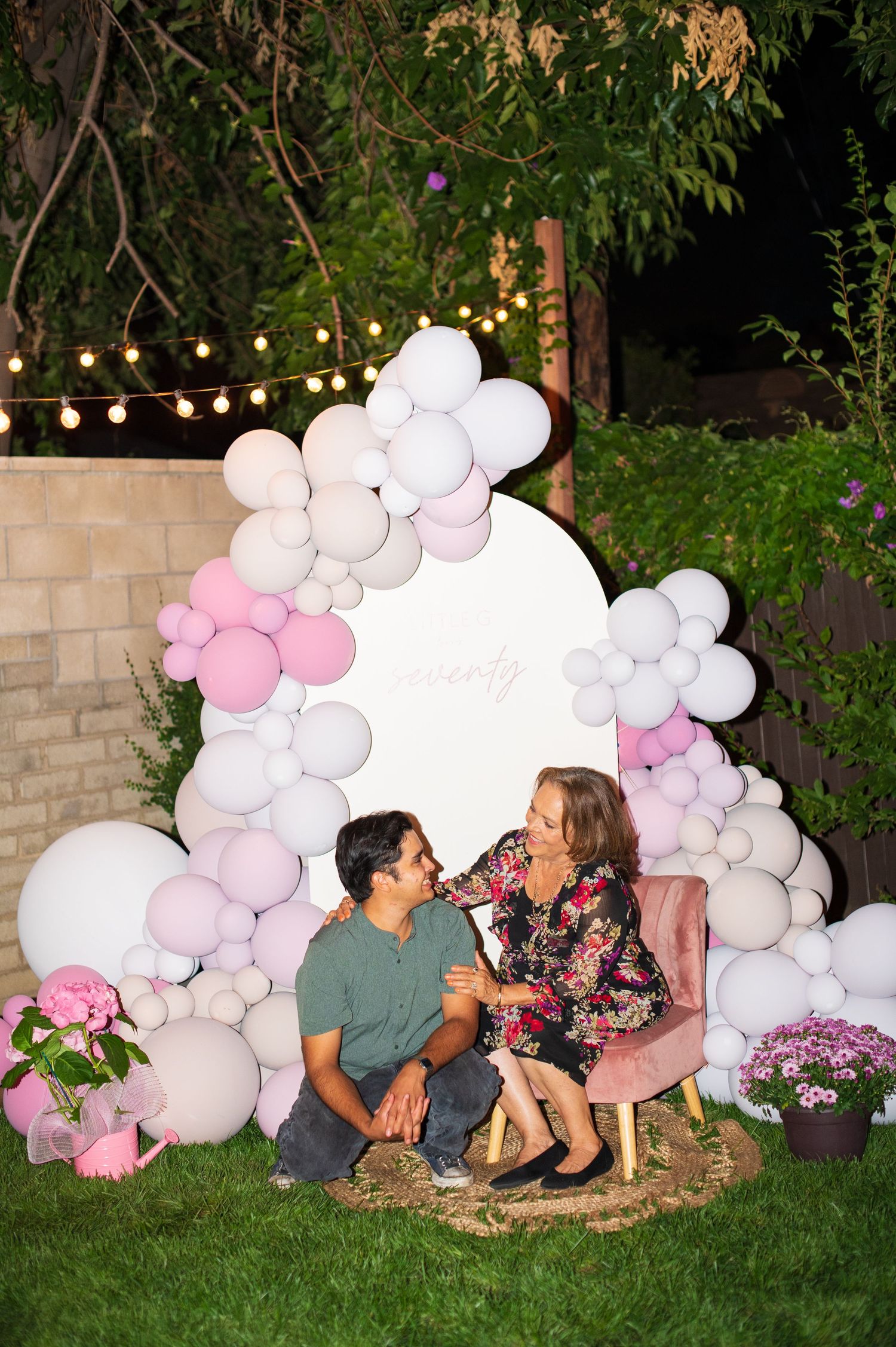 An outdoor evening celebration scene with string lights and a decorative pink and white balloon arch installation.