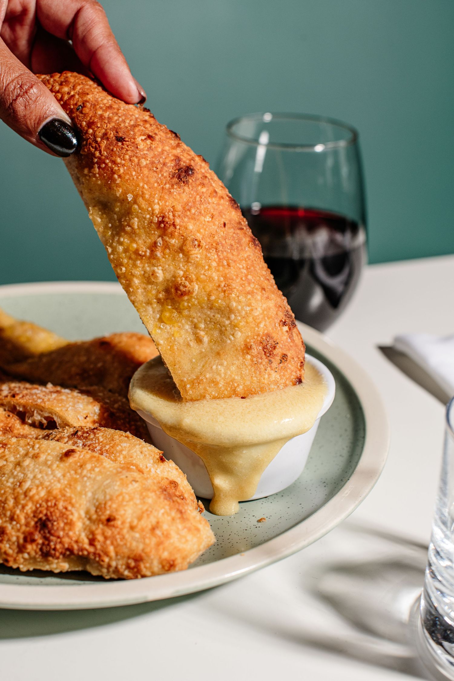 Hand dipping golden fried appetizer into creamy sauce with glass of red wine in background.