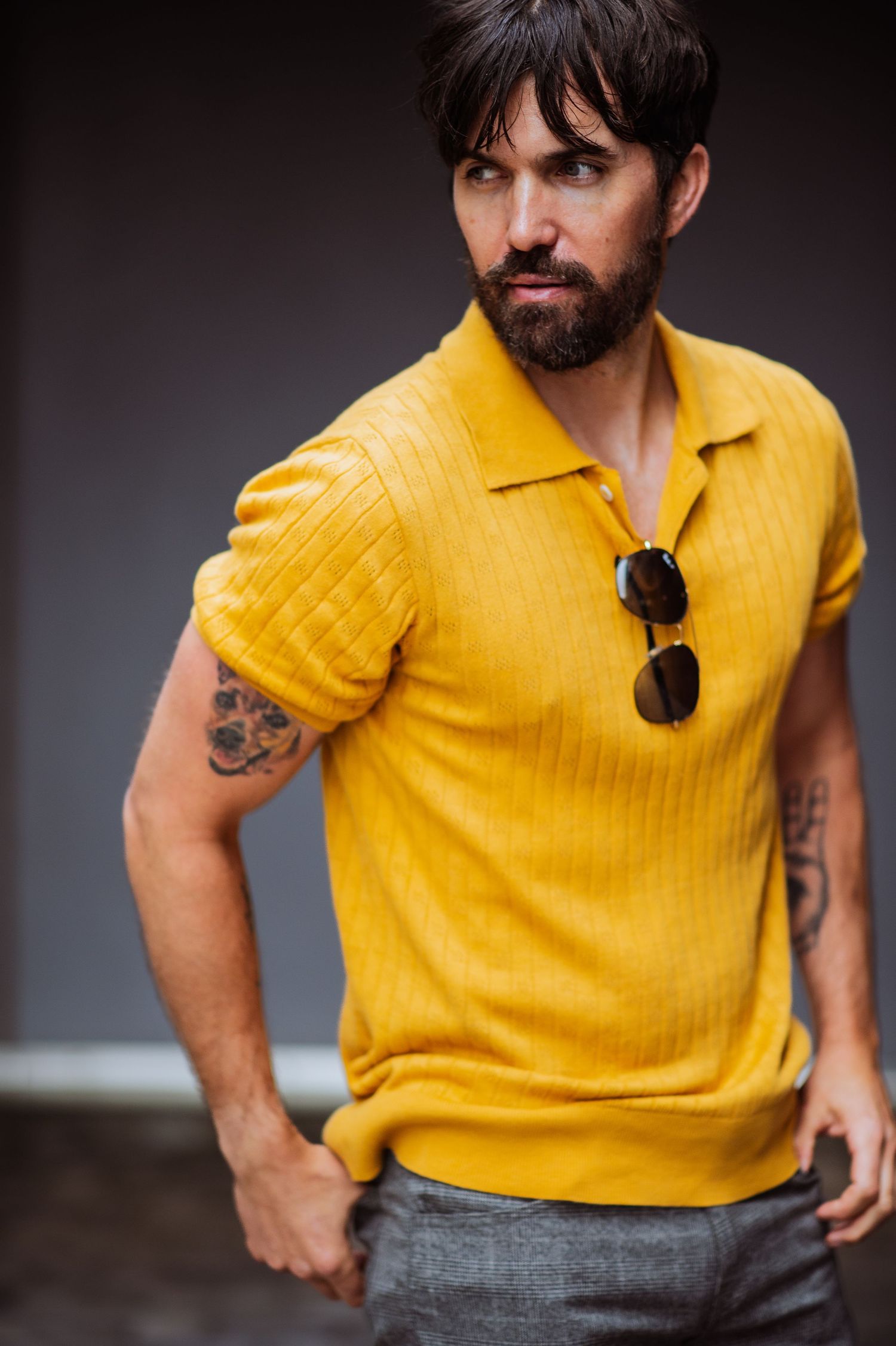 Bearded person wearing bright yellow polo shirt and sunglasses carries confident posture against dark background.