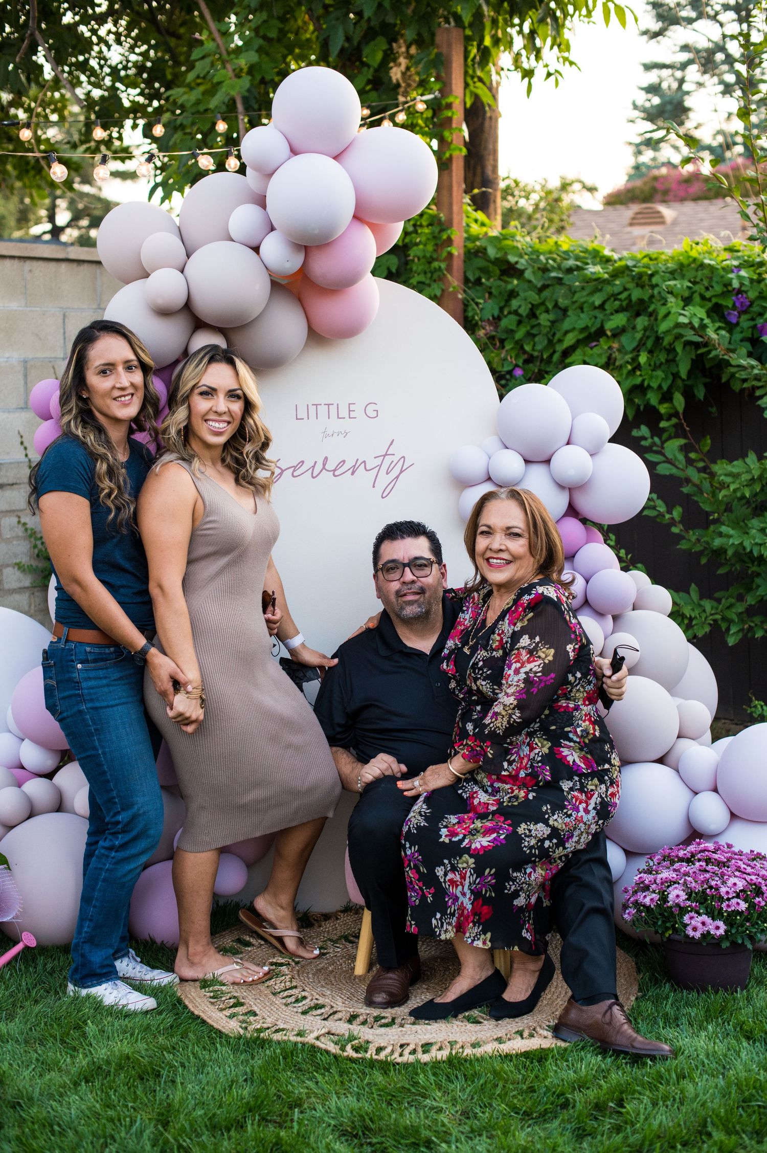 Outdoor celebration setup with elegant balloon arch in white and pink tones alongside floral decorations in a garden setting.
