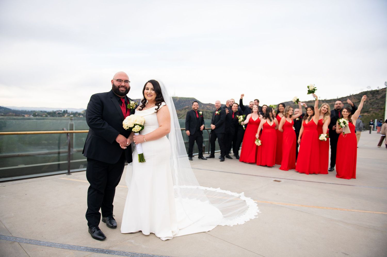 Wedding party members in red dresses and formal attire pose together on a scenic outdoor walkway.