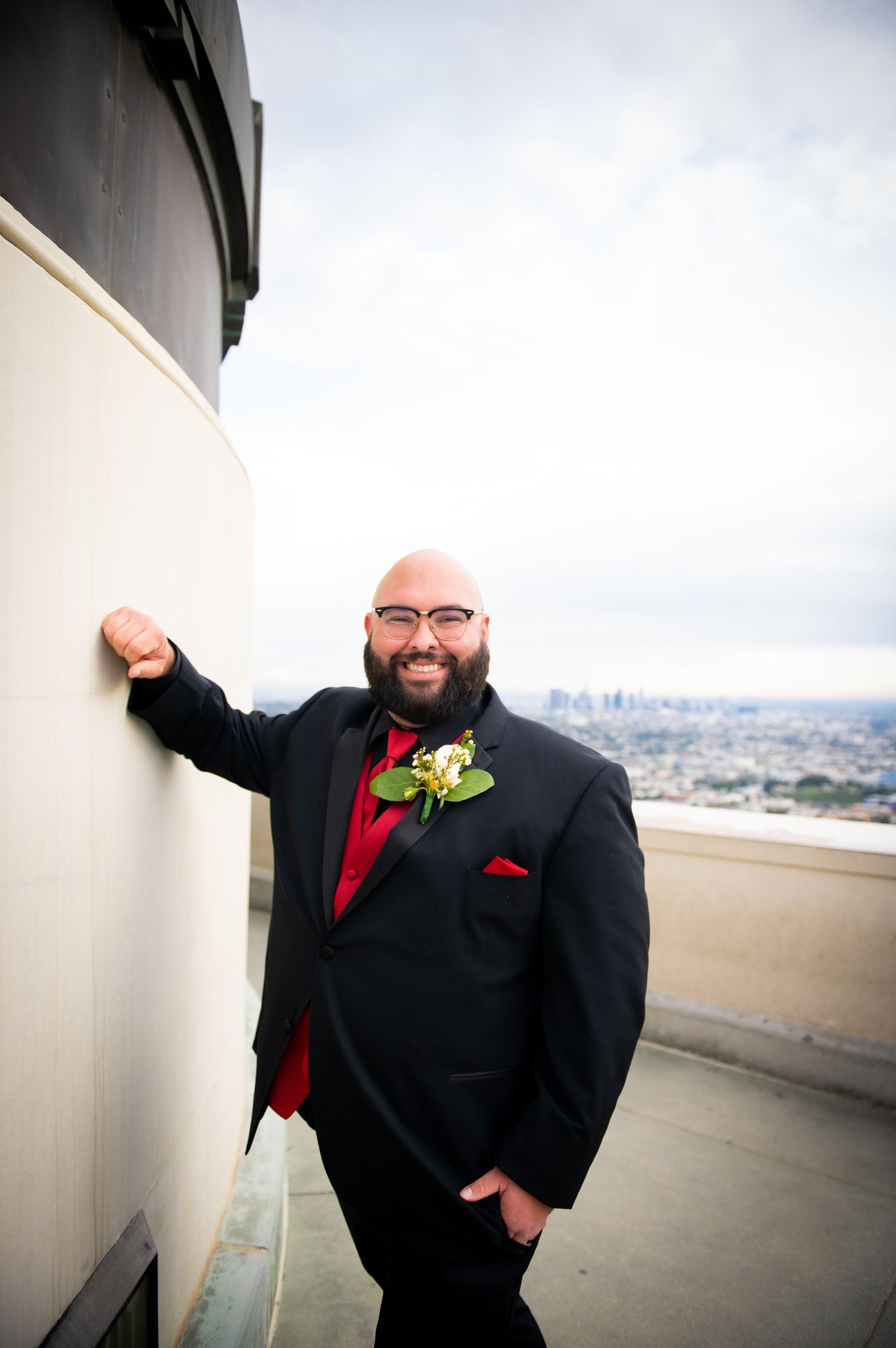 A groom in a black suit with red accents and boutonniere poses against a curved wall on a rooftop venue.