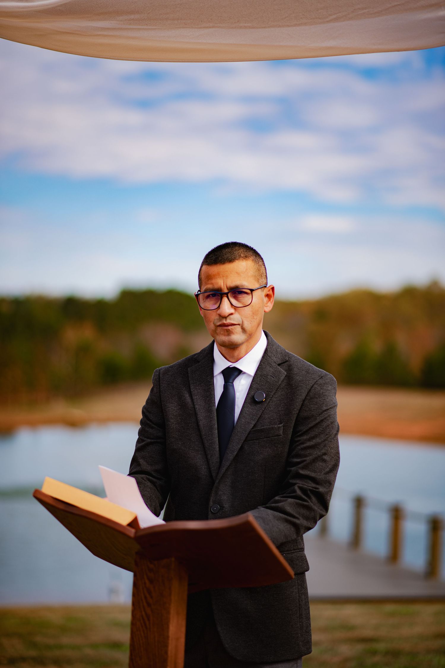 A person in a black suit and glasses reads from a large book at a wooden podium by a serene lakeside.