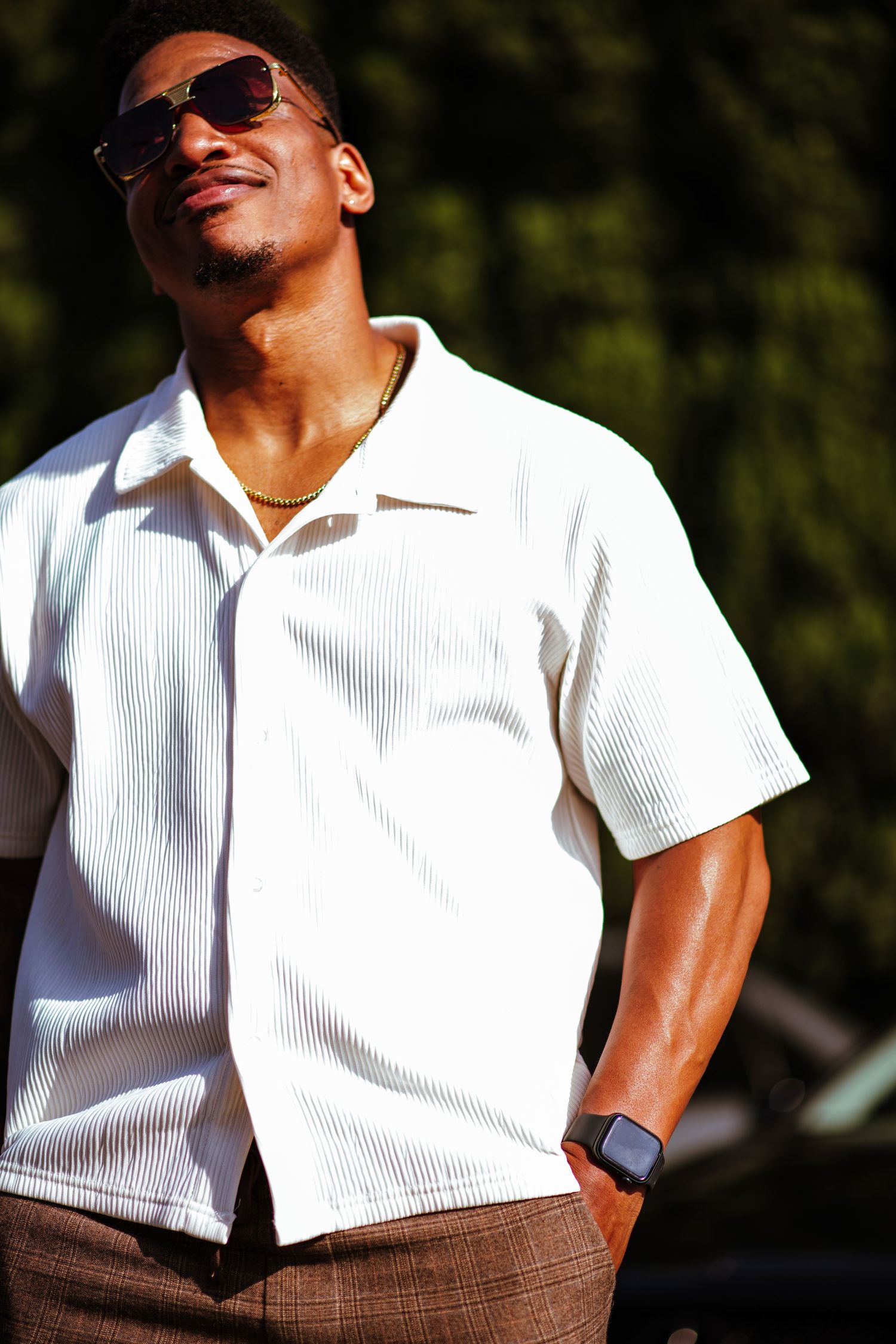 Person in white polo shirt and brown pants poses casually in natural outdoor lighting.