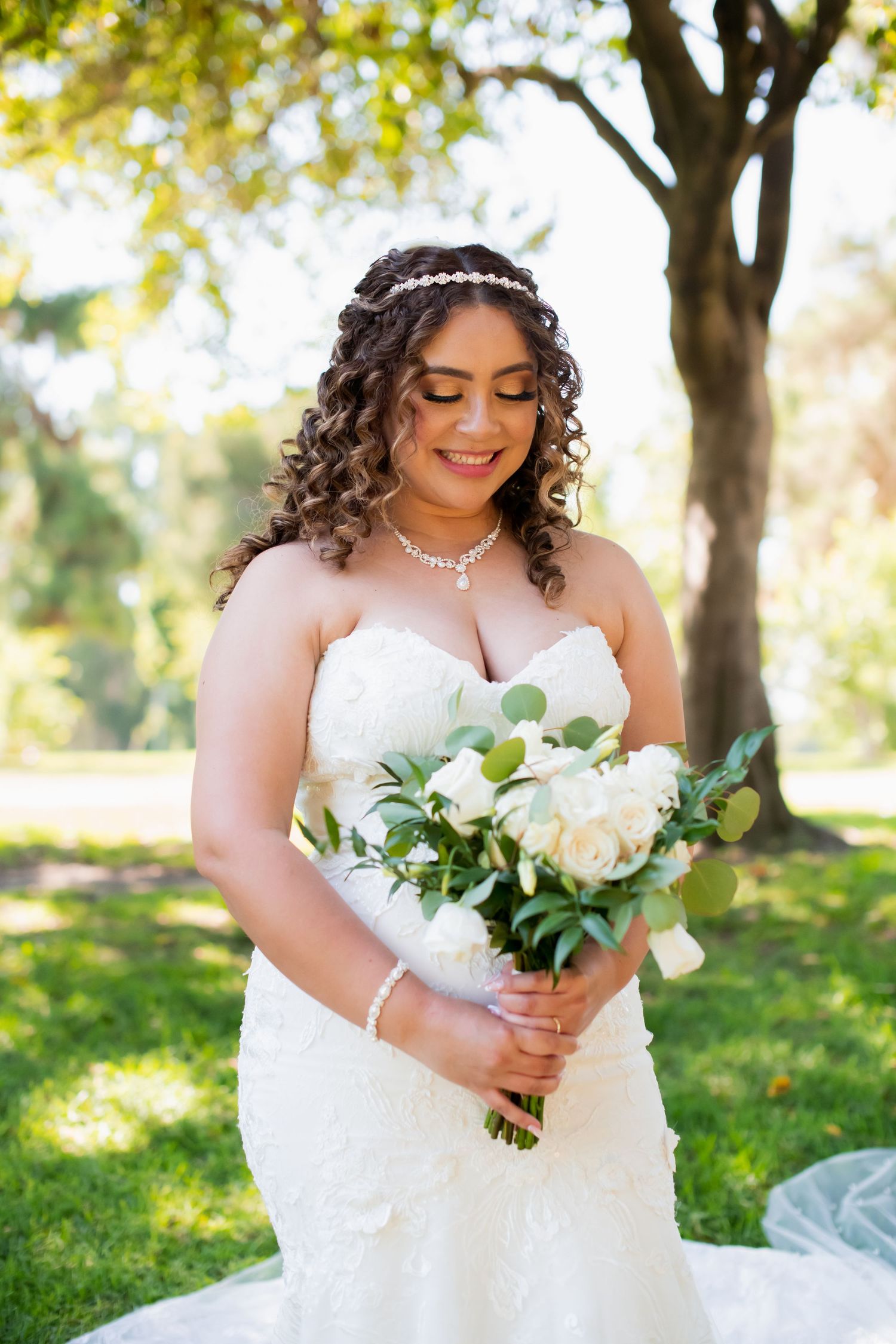 Bride with curled hair holds white and green bouquet while smiling downward.