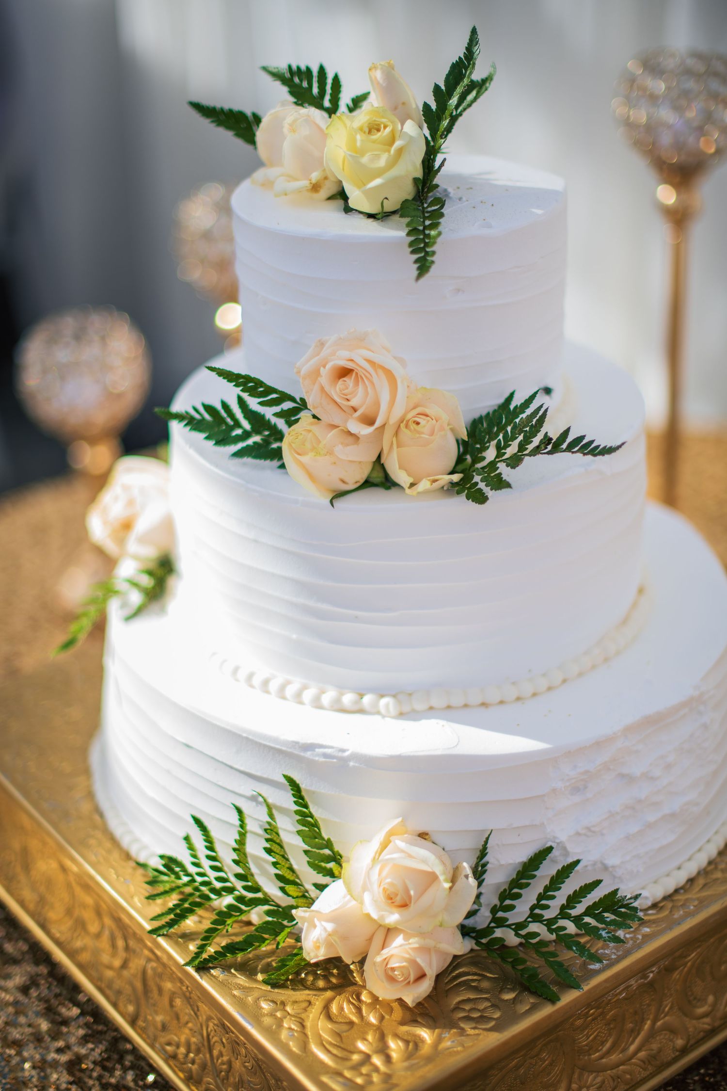 Classic white wedding cake decorated with peach roses and green ferns on gold stand.