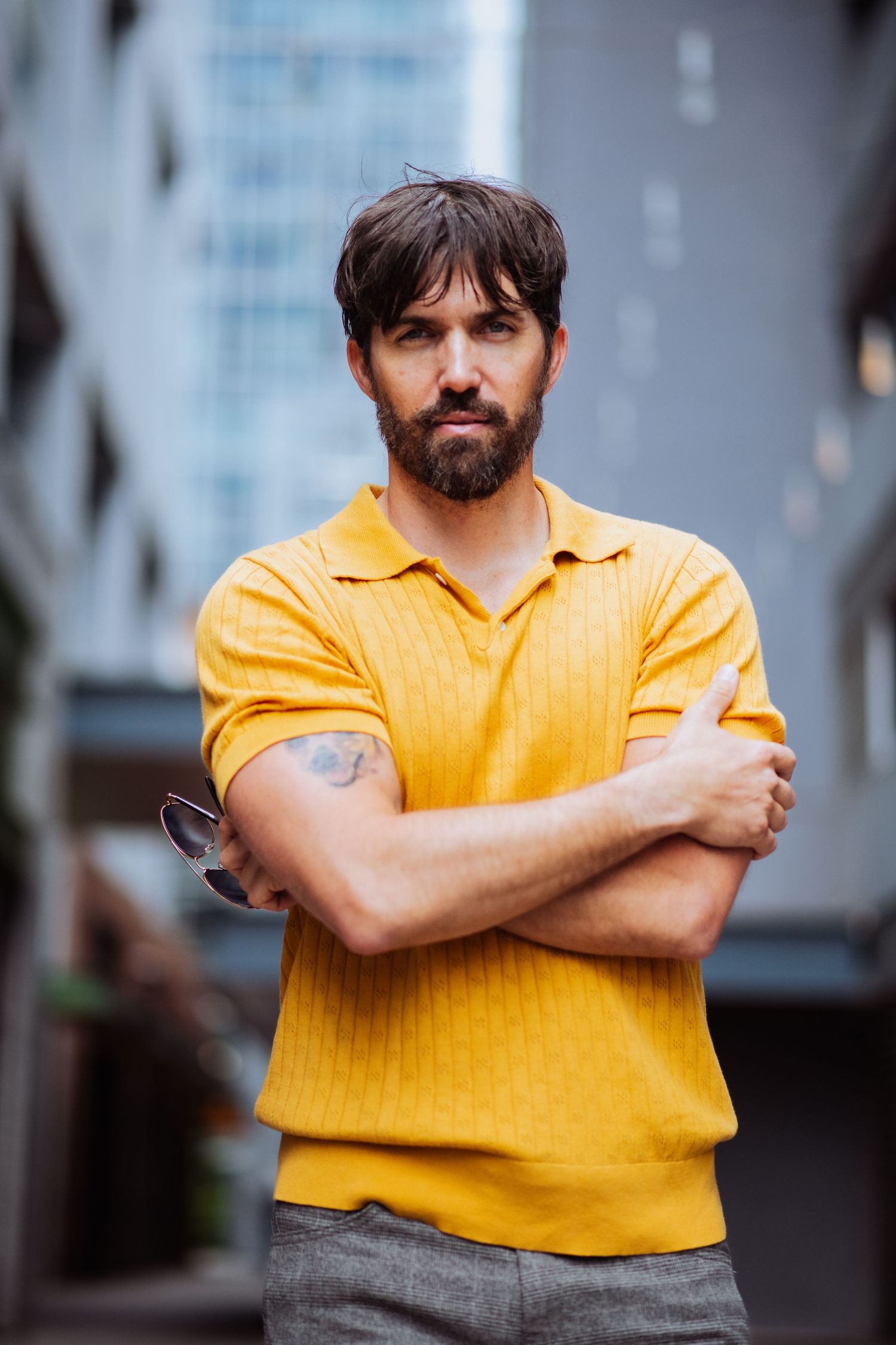 Figure in bright yellow shirt crosses arms while posing outdoors with urban background.