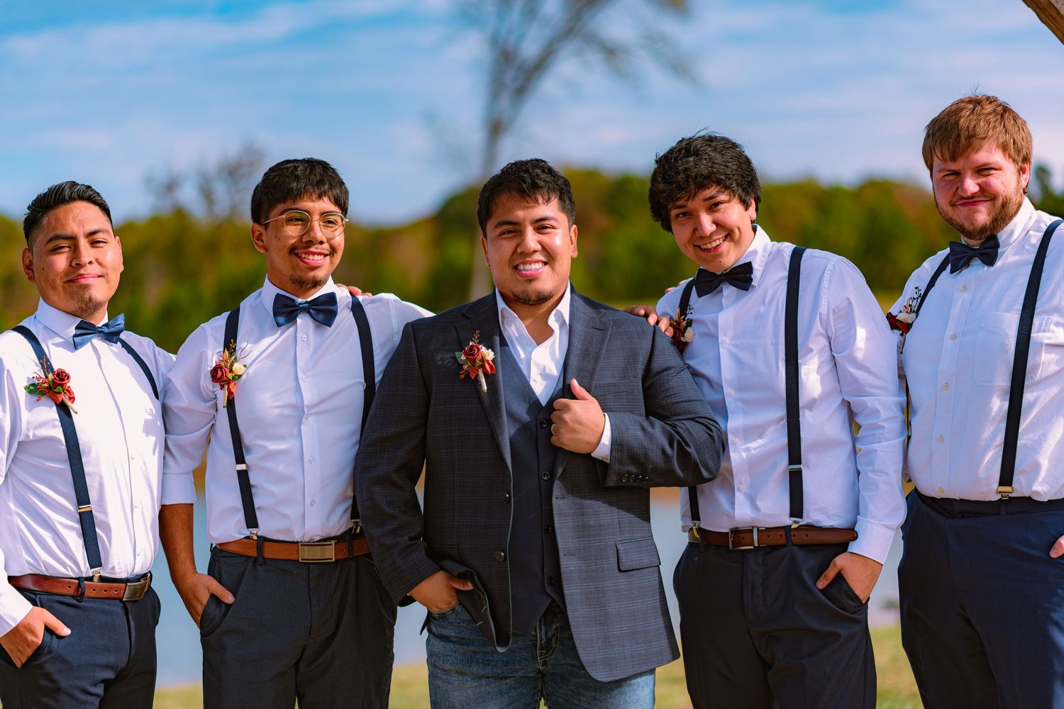 A group of five groomsmen in formal attire with suspenders and bowties pose together outdoors on a sunny day.