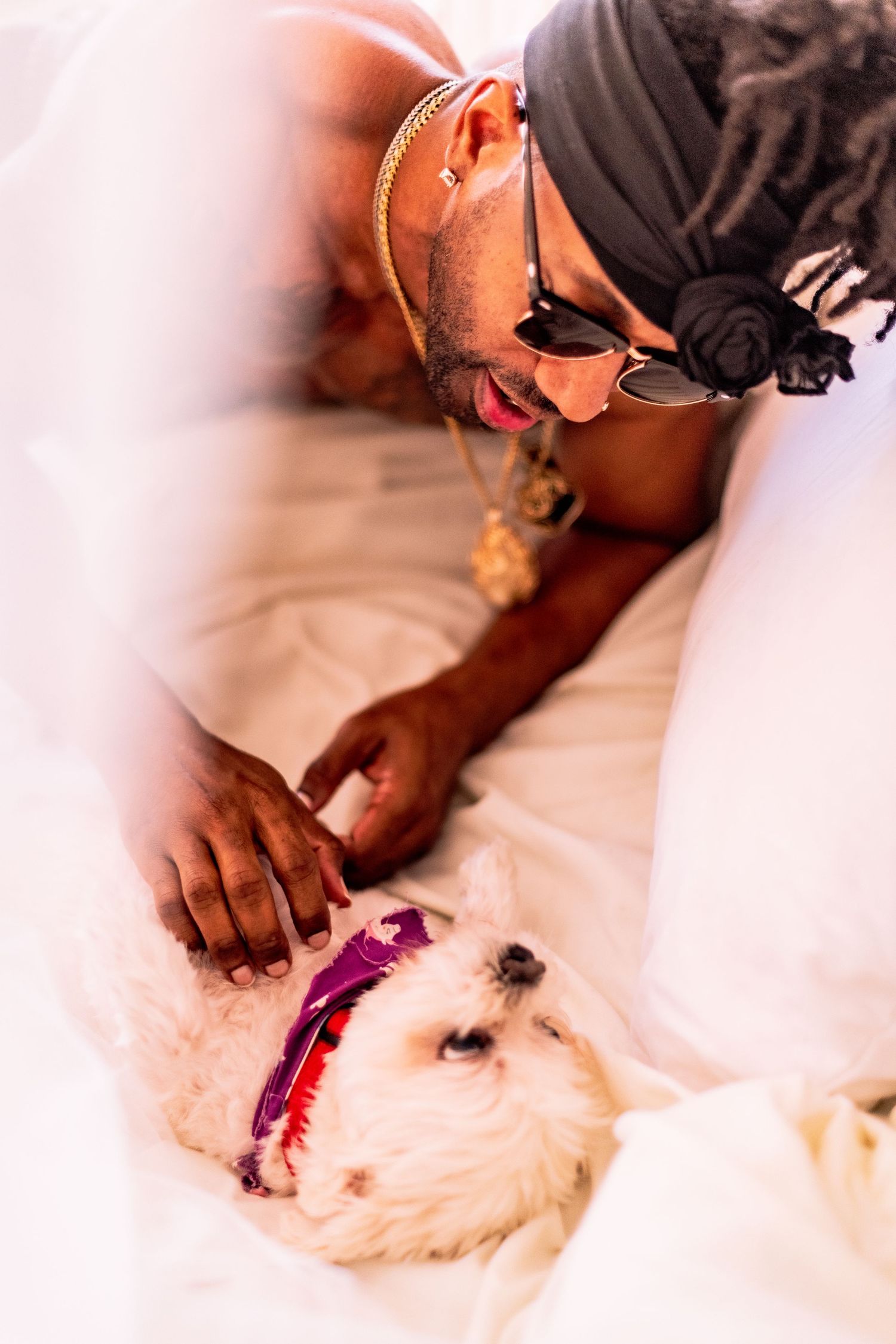 LongLiveCzar wearing sunglasses and jewelry pets a small white dog Coop Devillington with a pink collar lying on white bedding.