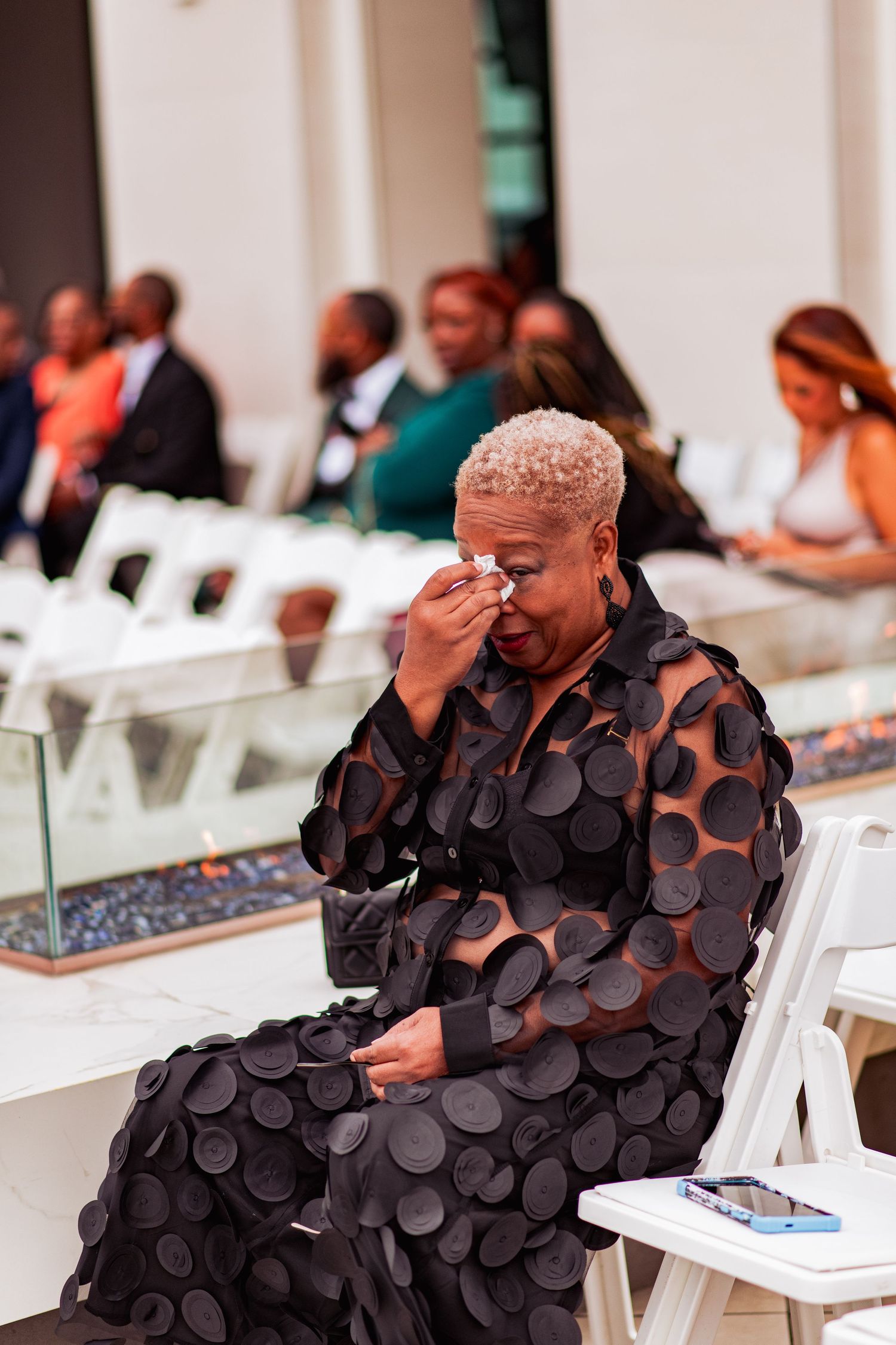 A person in a textured black dress sits thoughtfully on a white chair during an event.