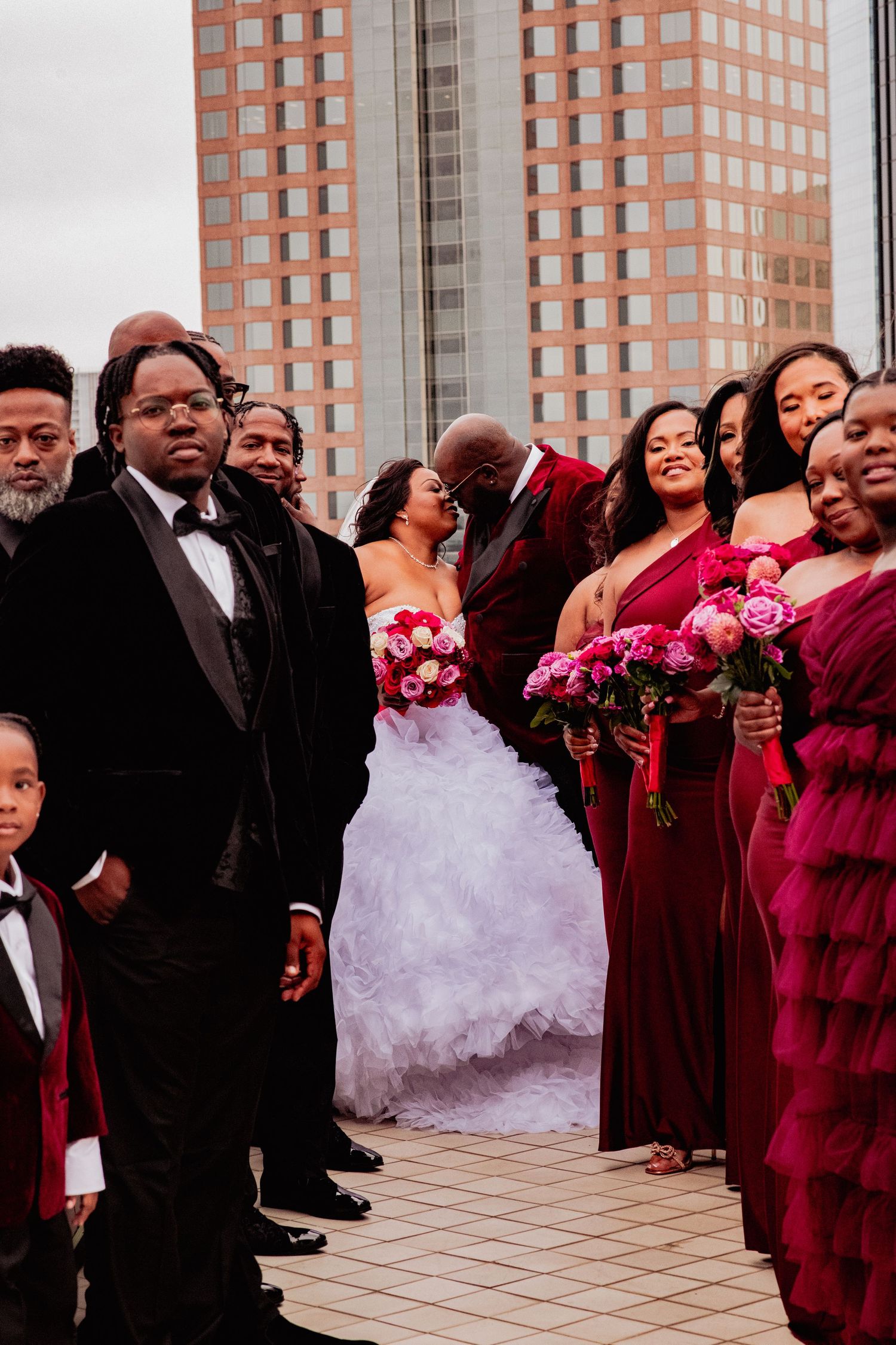 Wedding party members in burgundy dresses and black suits gather on a terrace with a bride in a white gown.