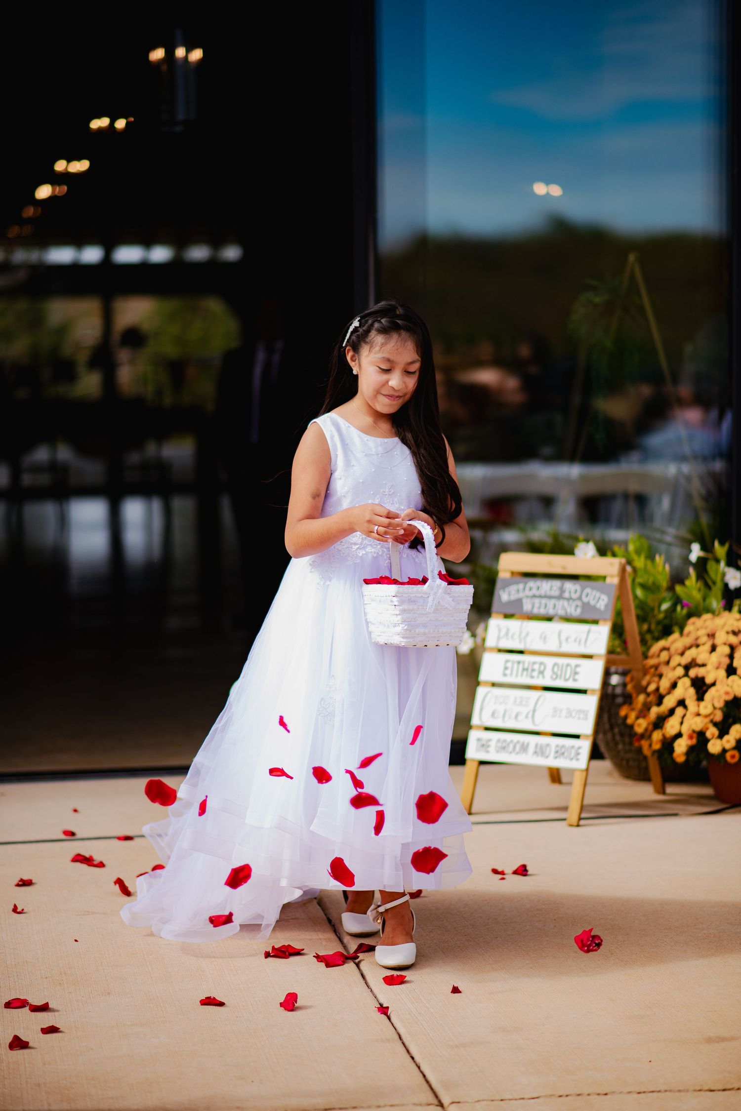 Young flower girl in white dress scatters rose petals on outdoor pathway.