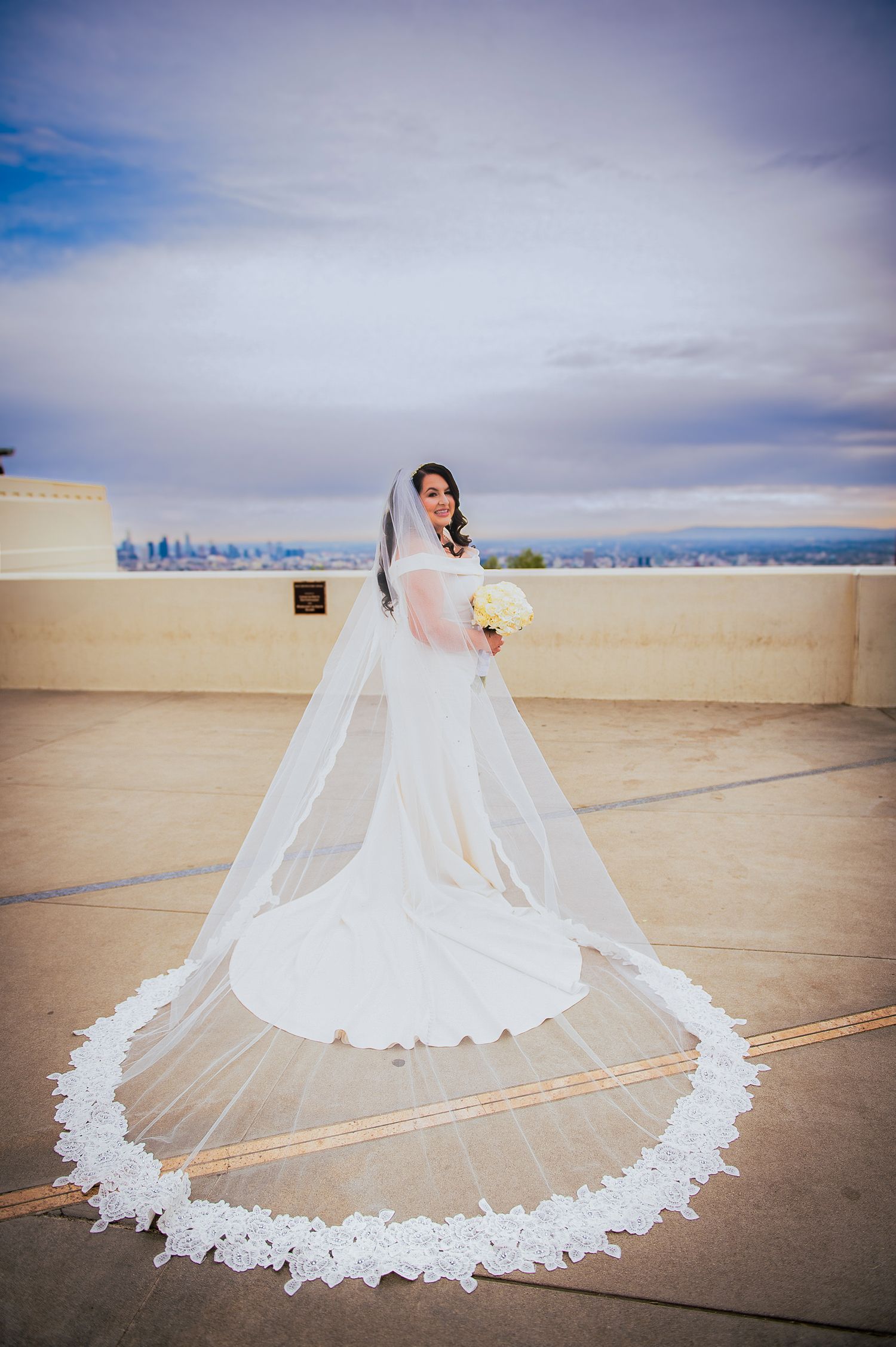 Dramatic cathedral-length lace wedding veil spread across concrete with city skyline in background.