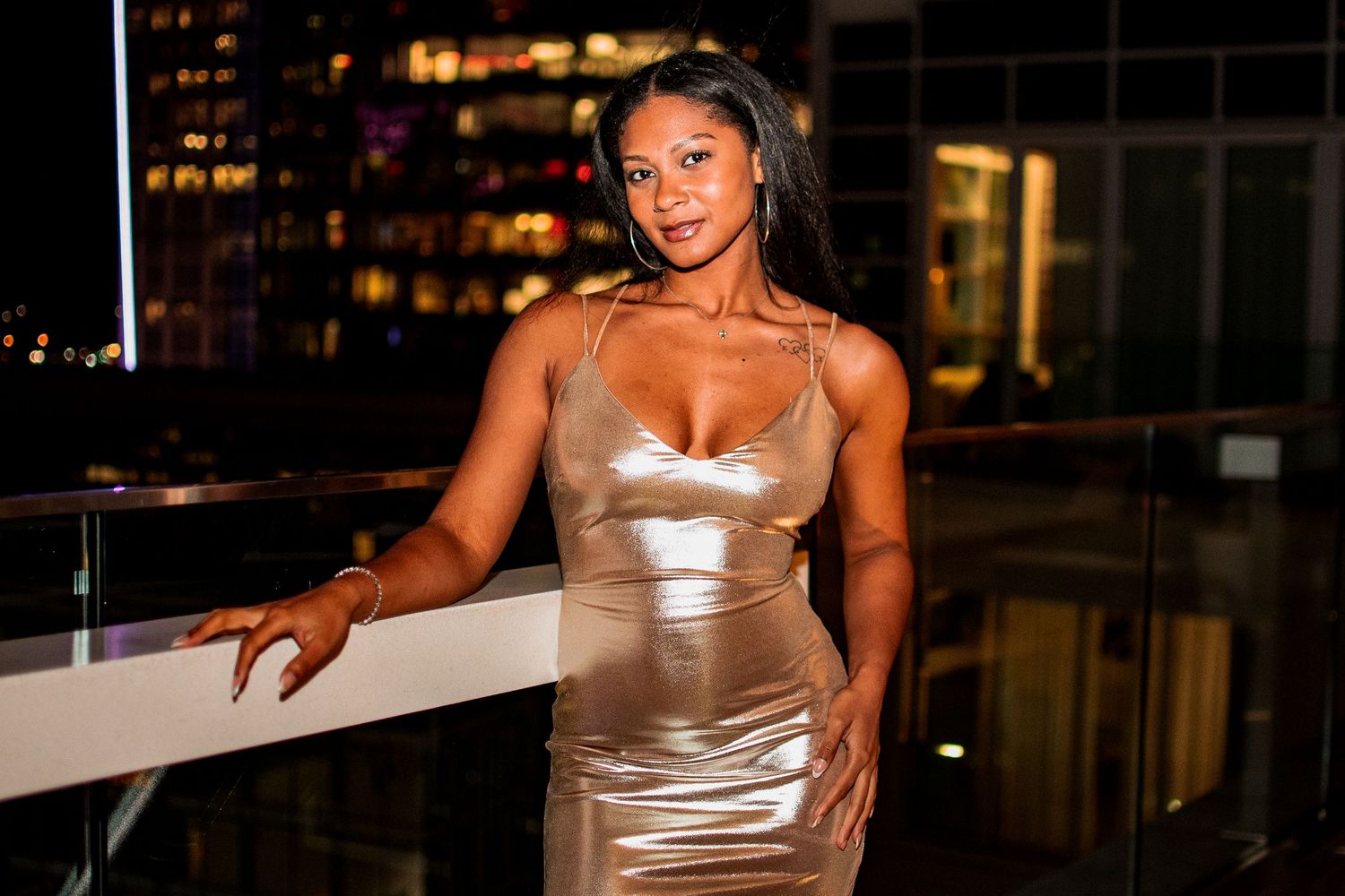 A woman in a champagne satin dress poses against a railing with city lights behind her.