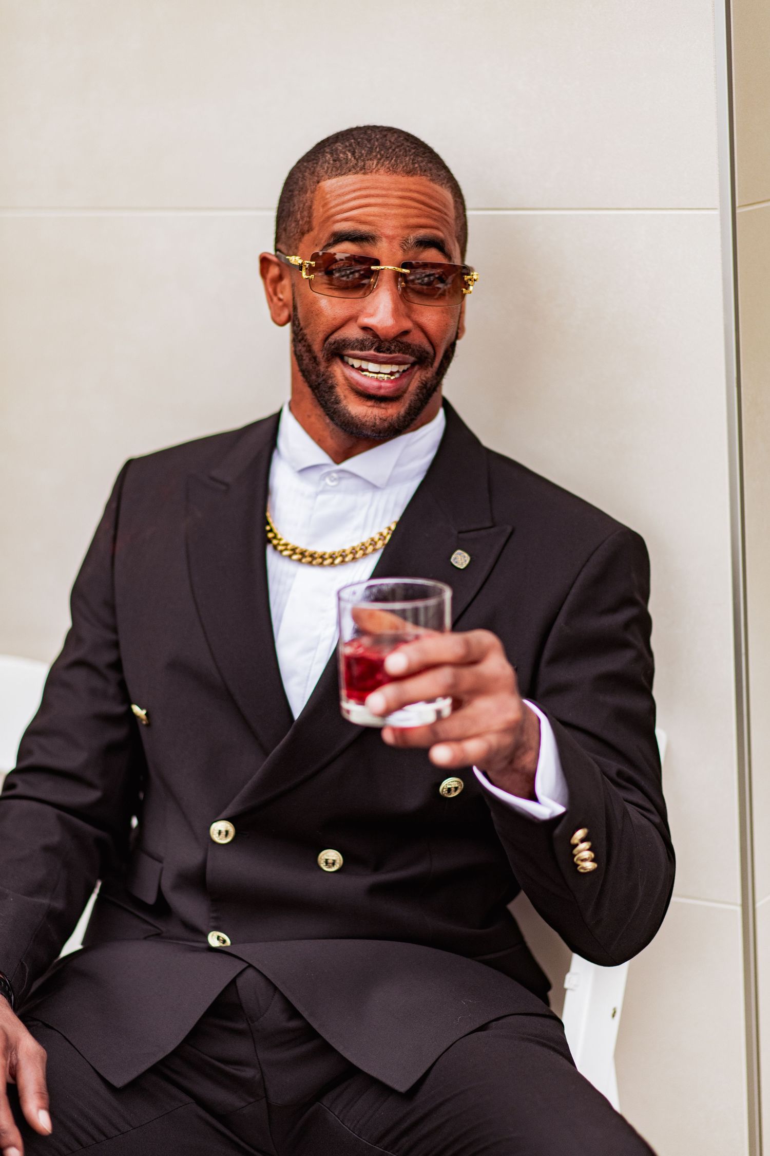 A person in a black suit and gold chain holds a drink while smiling during a celebration.