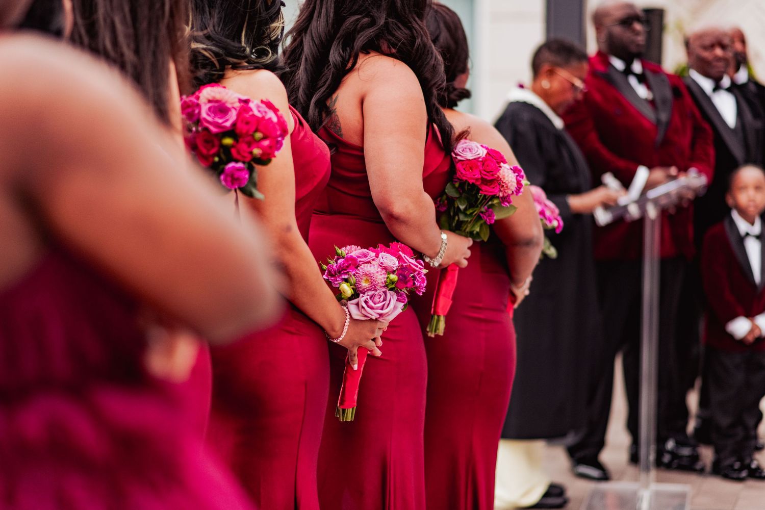Wedding party members hold bright pink bouquets while wearing deep burgundy gowns.