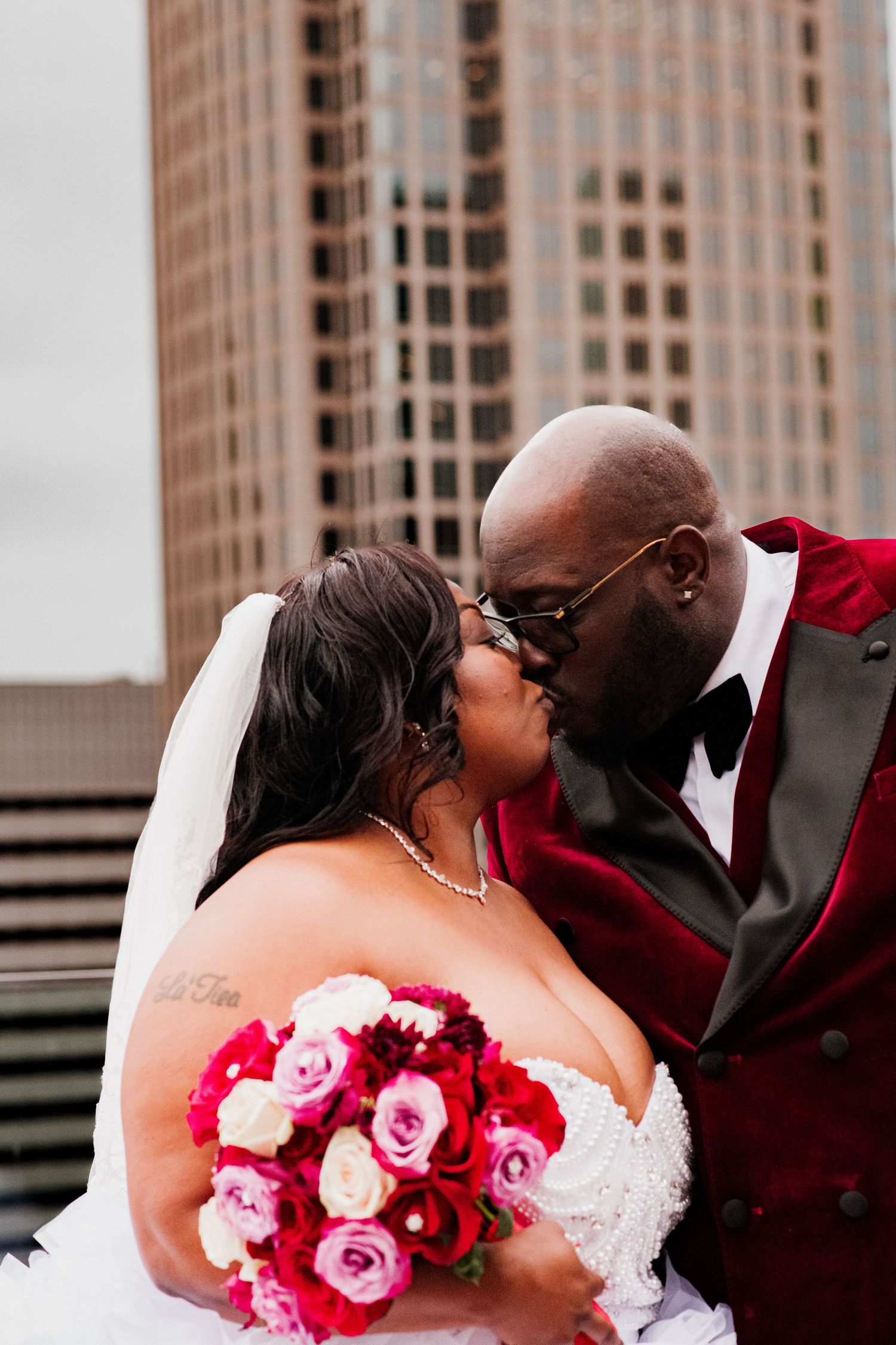 Wedding couple shares a romantic kiss against urban backdrop with pink and red rose bouquet.