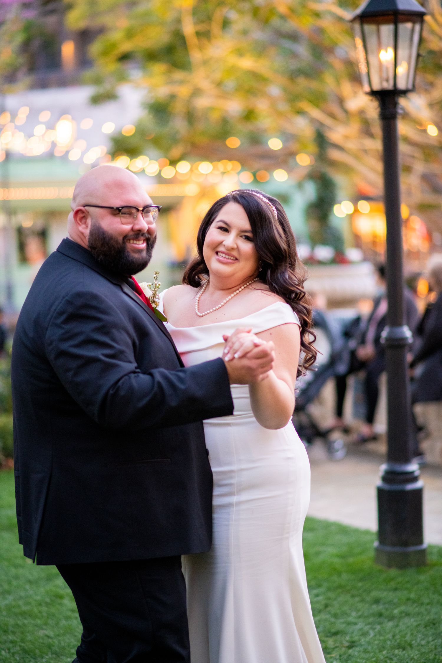 Newlyweds share first dance outdoors at twilight under string lights and vintage lamp post.