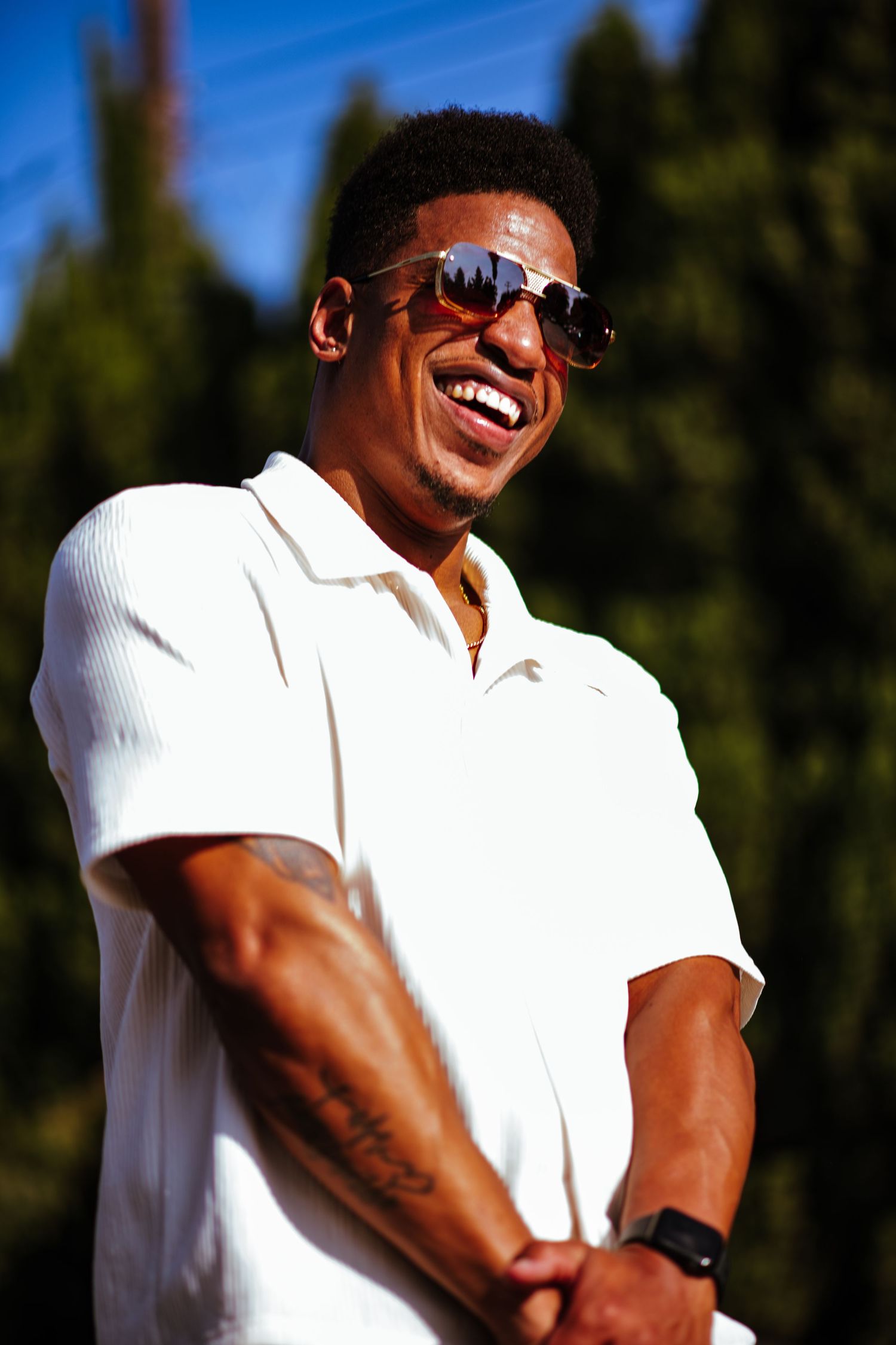 Person in white polo shirt wearing sunglasses smiles outdoors against green trees in bright sunlight.