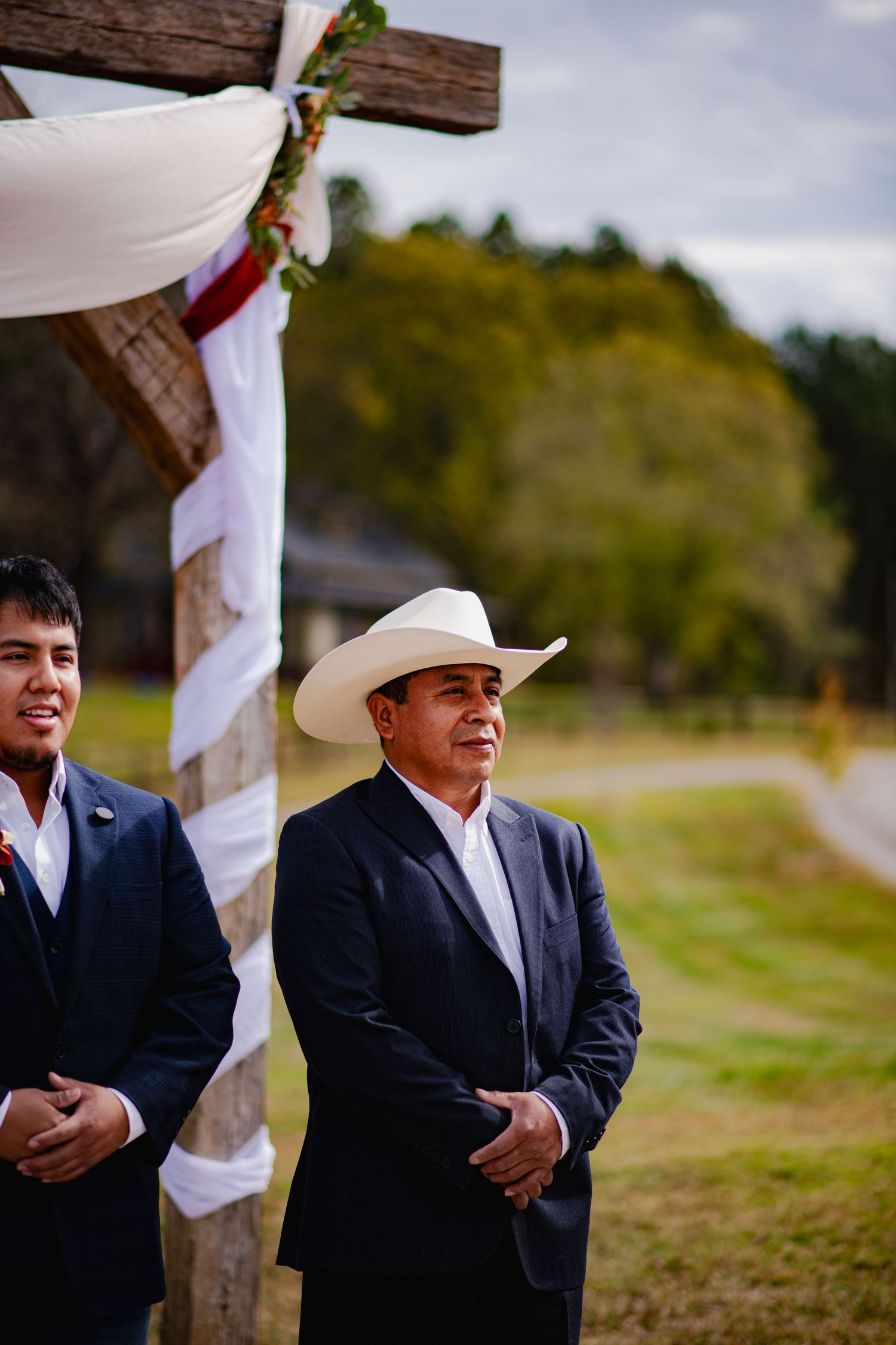 Wedding party members gather under rustic wooden arch decorated with white fabric during outdoor ceremony.