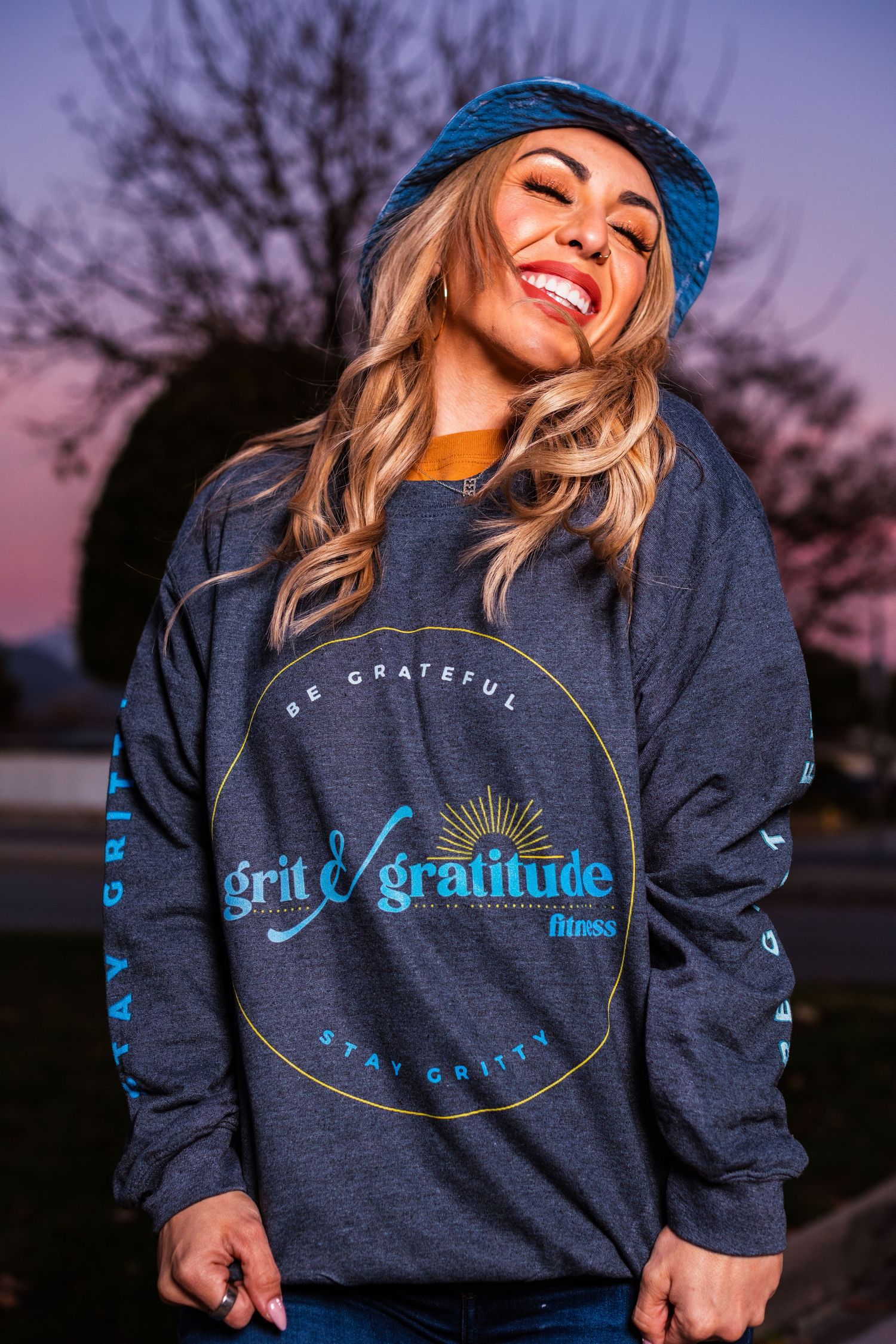 Dark gray sweatshirt with Grit and Gratitude logo worn with blue bucket hat at twilight.
