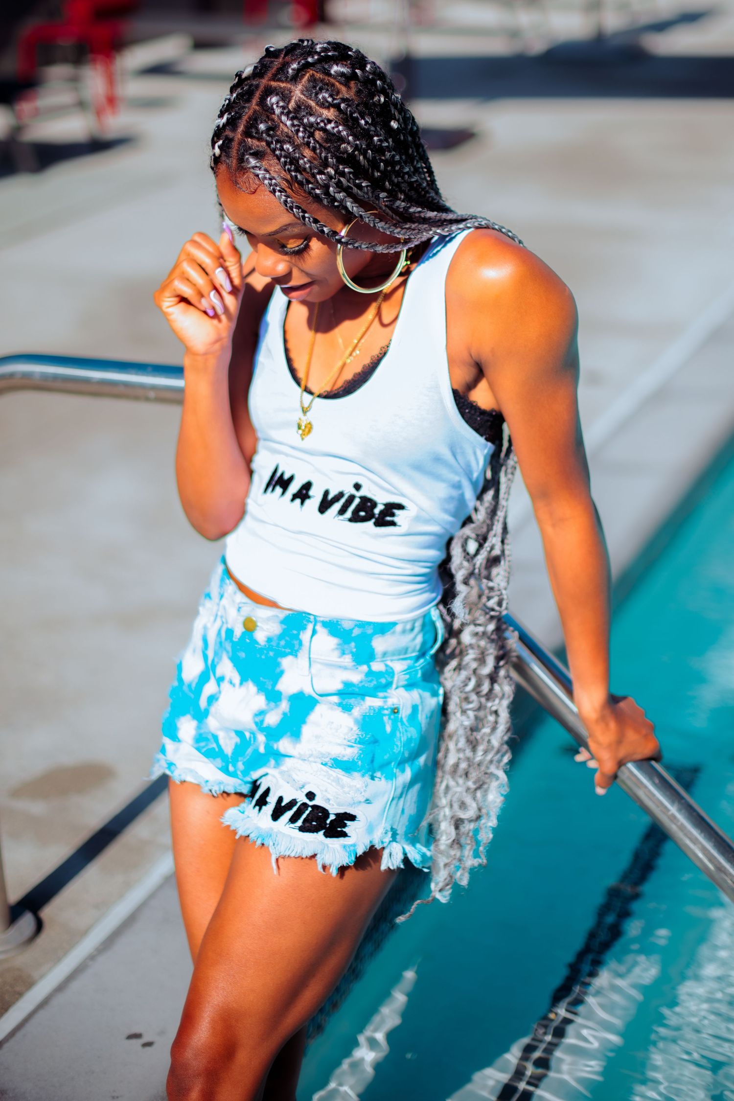 Model wearing tie-dye blue shorts and white tank top poses near railing in urban setting.