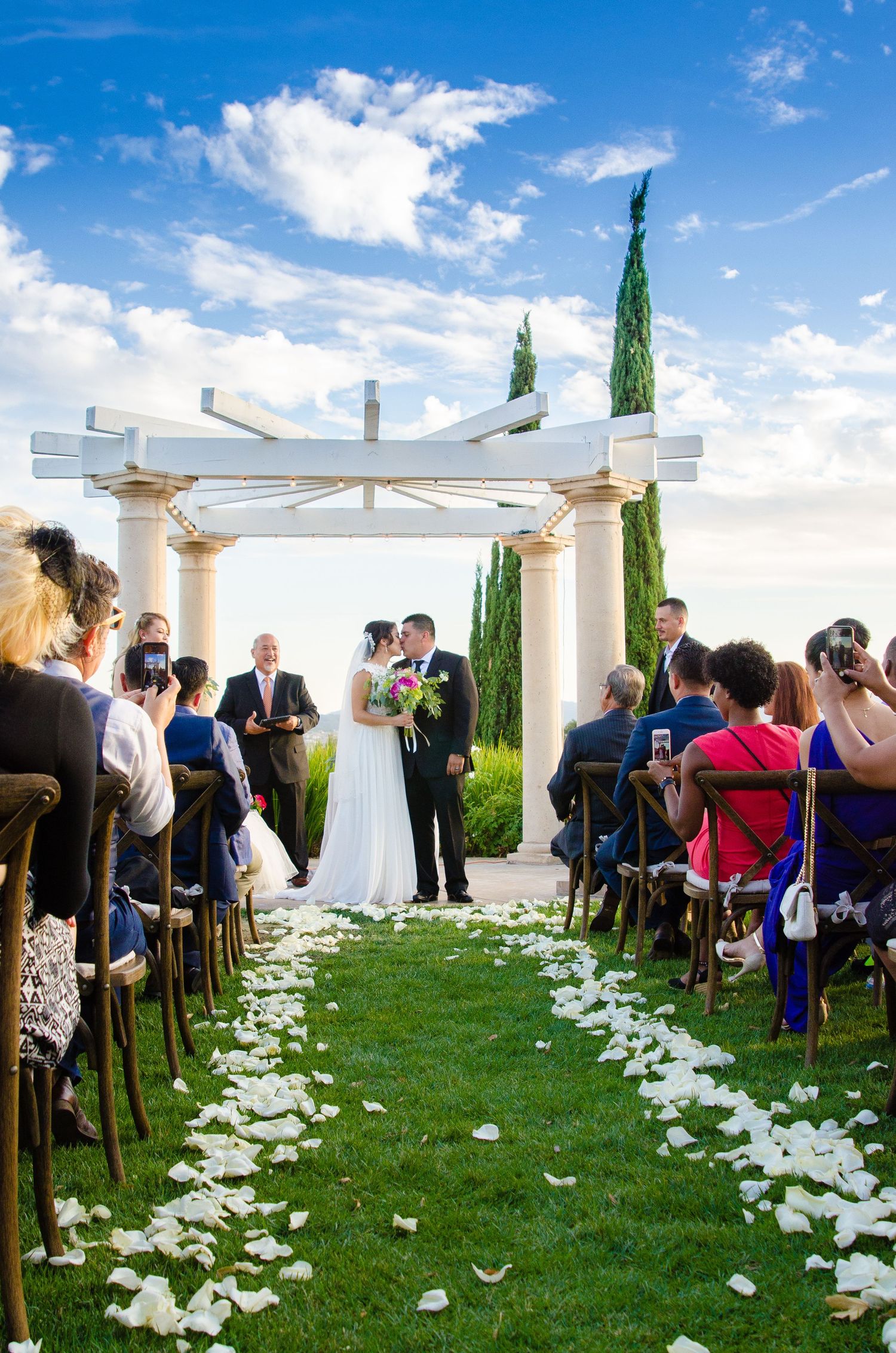 Outdoor wedding ceremony setup with white petals lining the aisle and white pergola under blue sky.