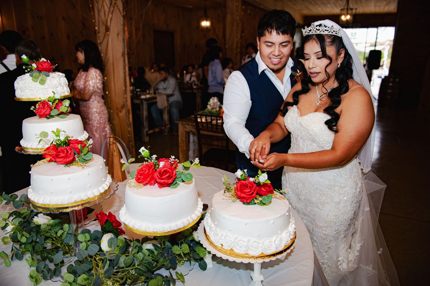 Wedding couple cuts into their tiered white cake decorated with red roses during reception.