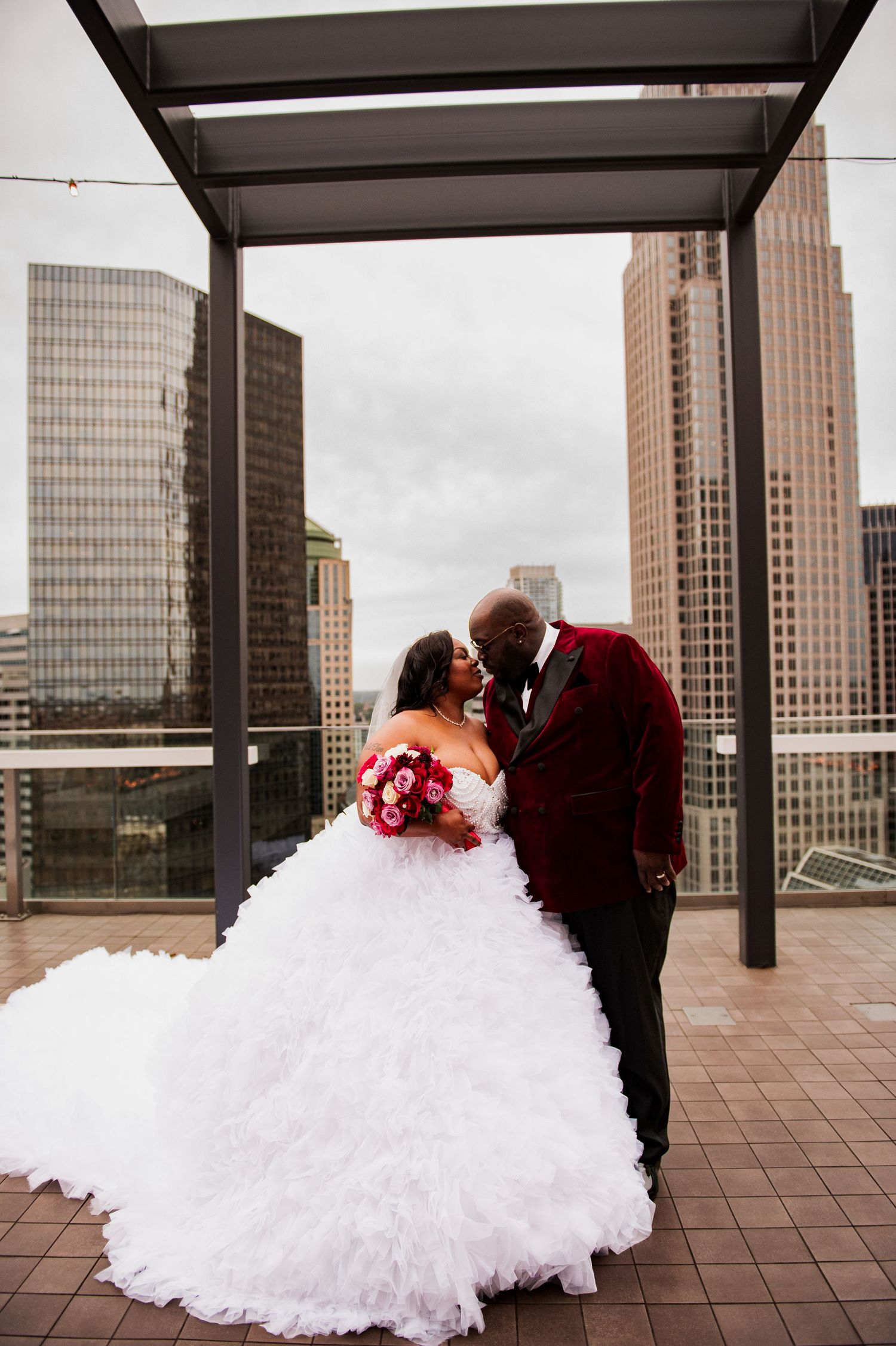 A romantic couple shares an intimate moment on a rooftop terrace with city skyscrapers in the background.