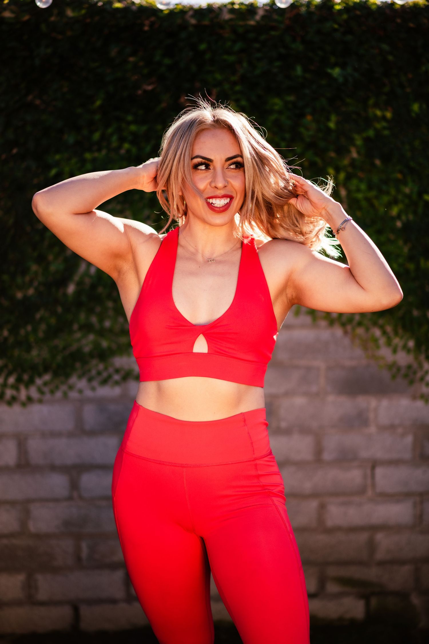 Athletic figure in vibrant red workout set poses confidently outdoors against a stone wall.