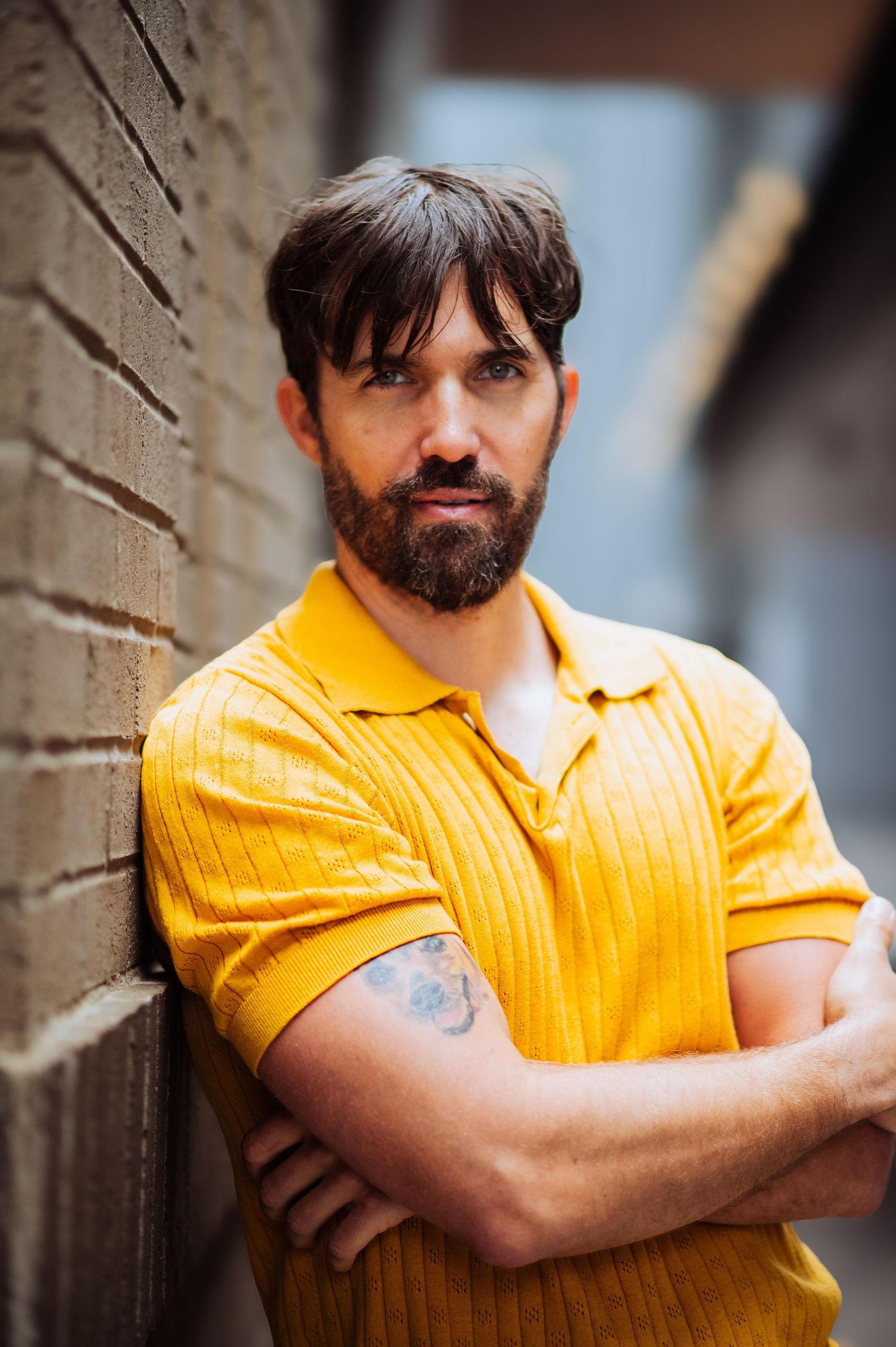 A casual portrait against a brick wall features a yellow polo shirt and relaxed pose.