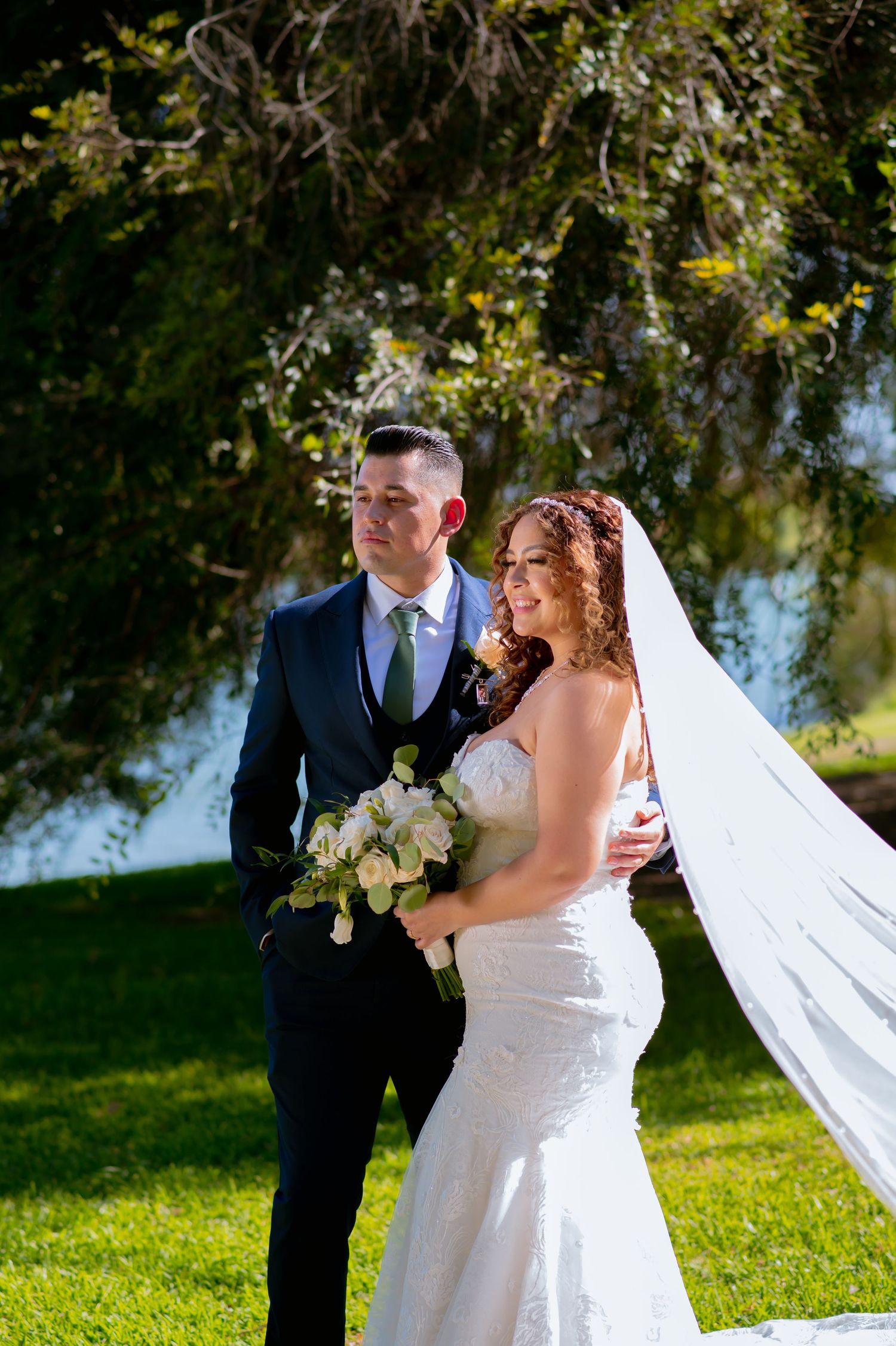 A romantic outdoor wedding scene with flowing veil against sunlit trees and greenery.