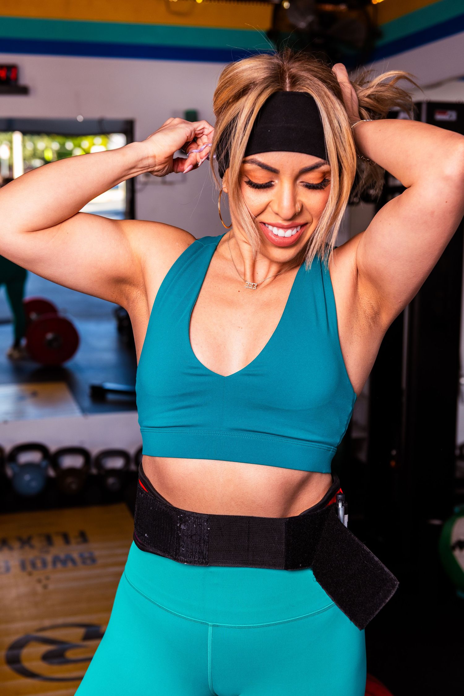 Woman in teal sports bra and black waistband exercising in a gym setting.