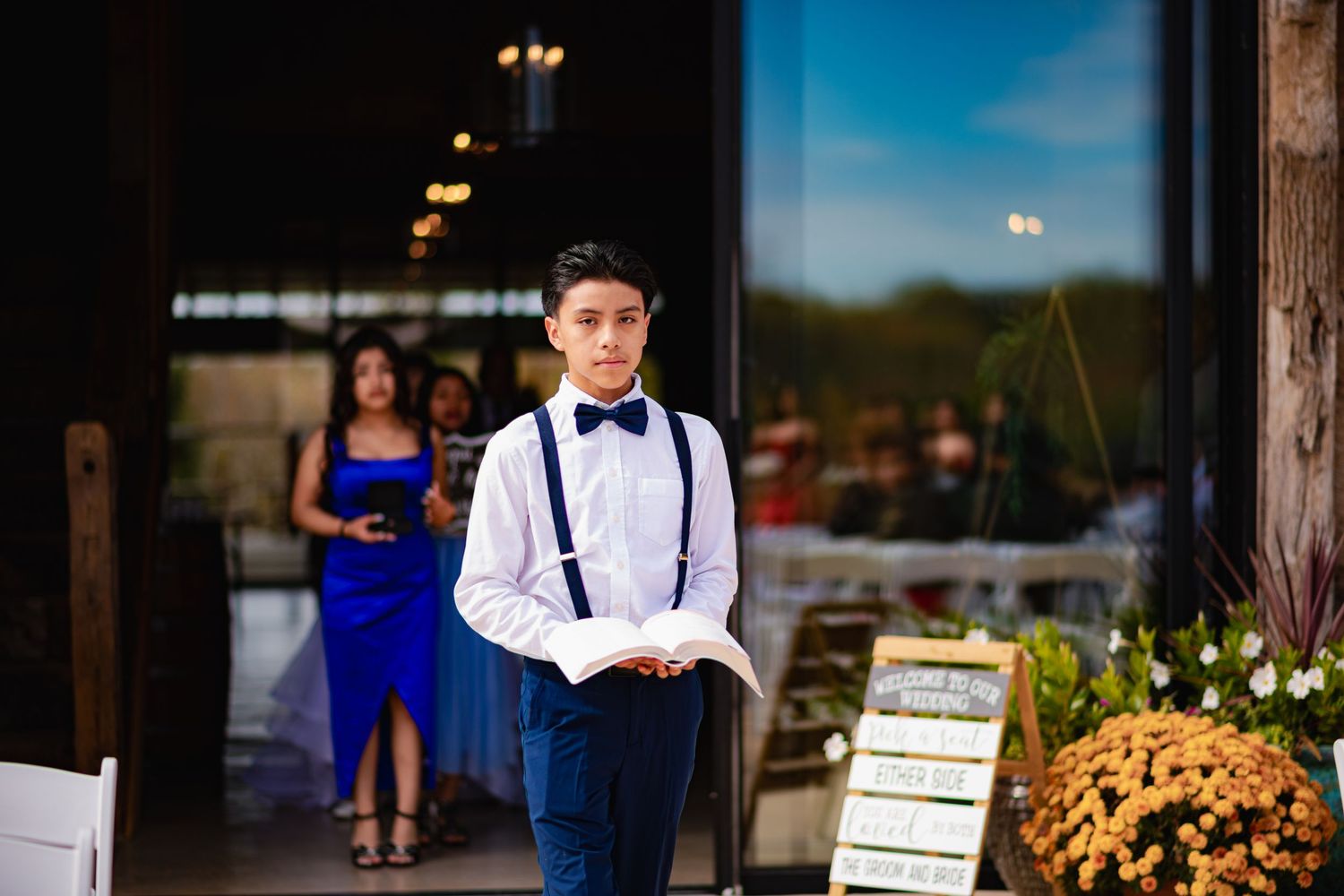 Someone in suspenders and bowtie stands in a rustic venue setting with decorative flowers and signage in the background.