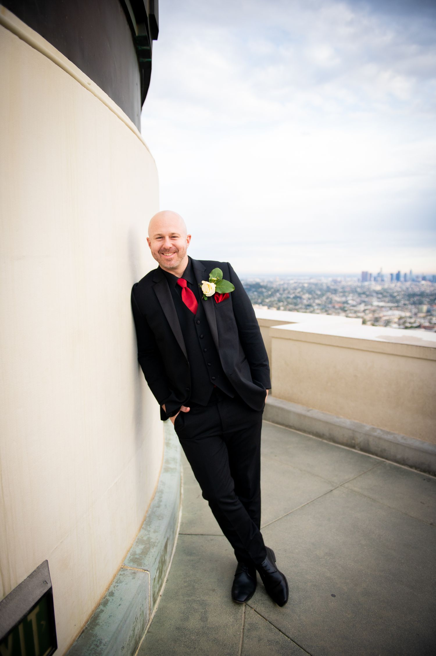 A groom in classic black tuxedo leans against a curved wall with city skyline visible in the background.