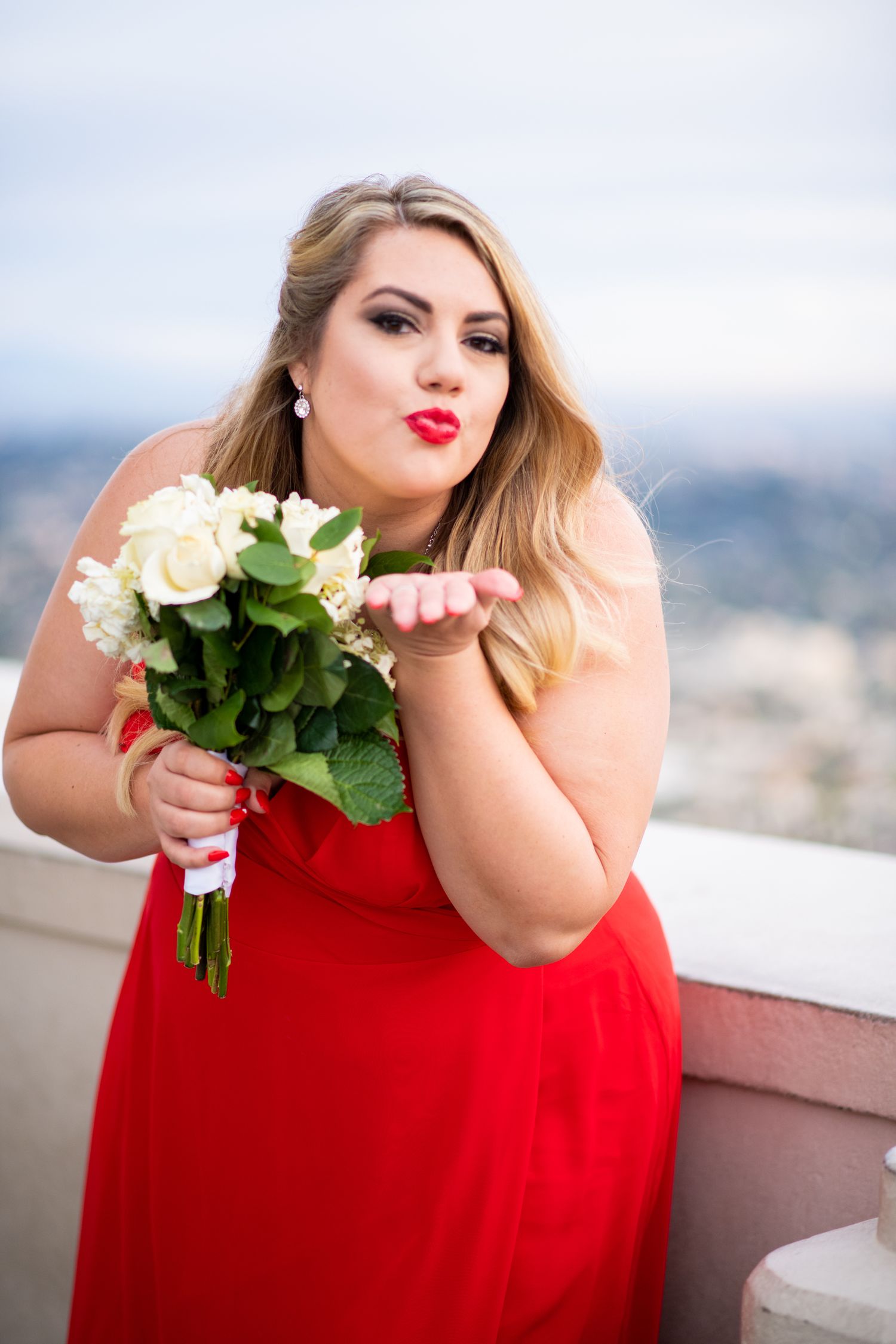 Woman in red dress blowing a kiss while holding white roses on a rooftop with city view in background.