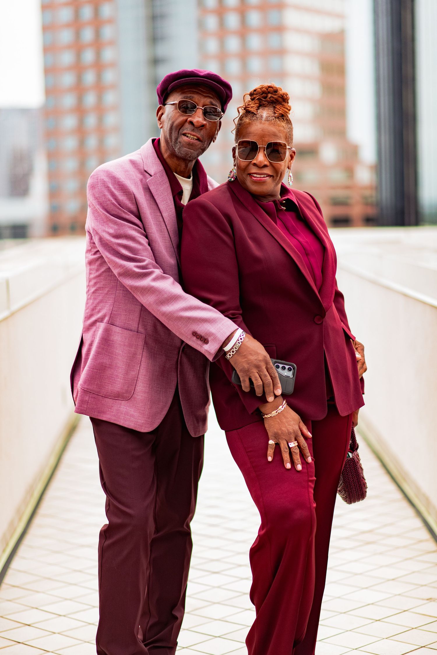 A stylish couple poses outdoors wearing coordinating pink and burgundy formal attire against an urban backdrop.