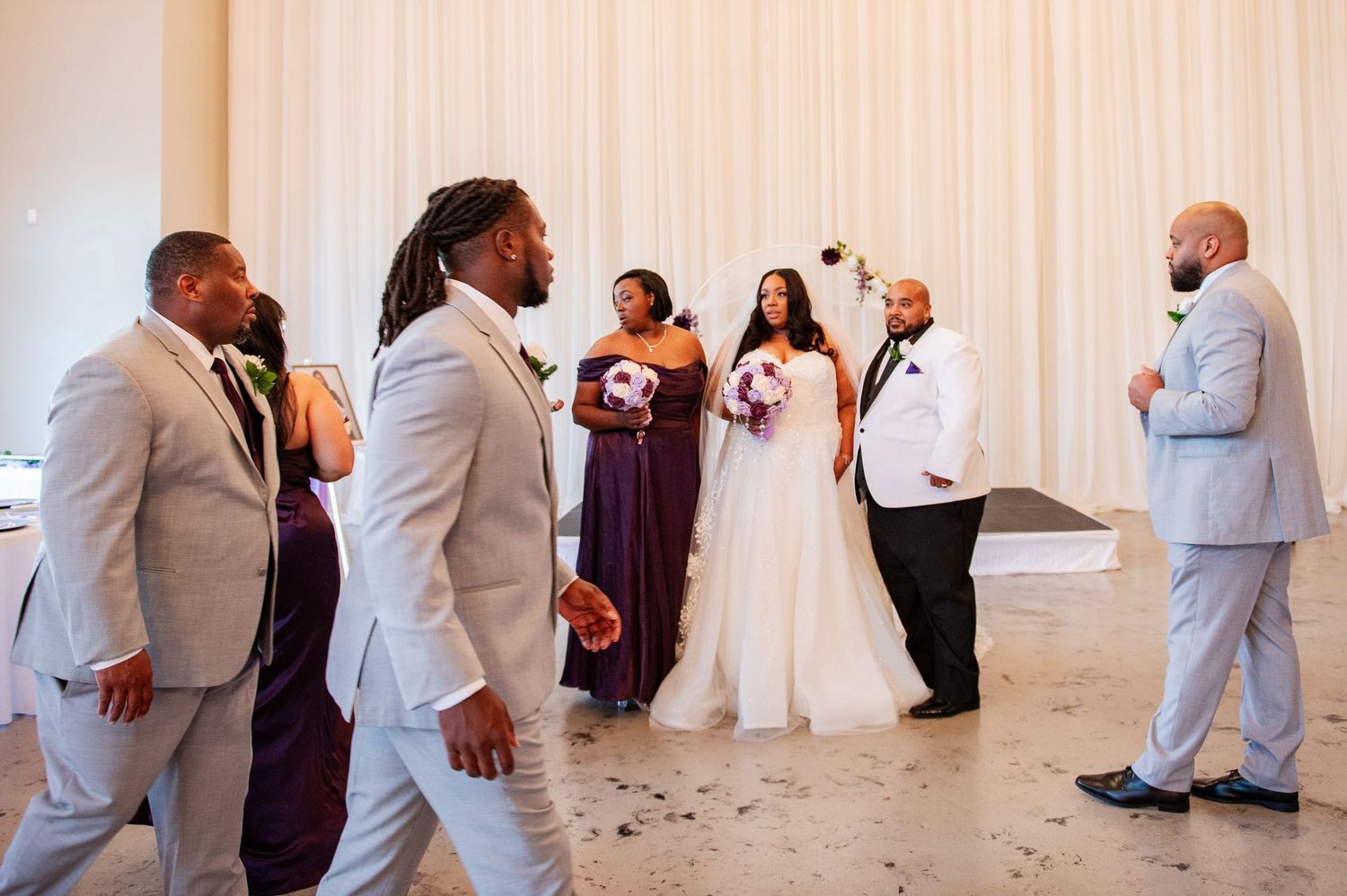 Wedding party members in light suits and purple dresses gathered in reception hall.