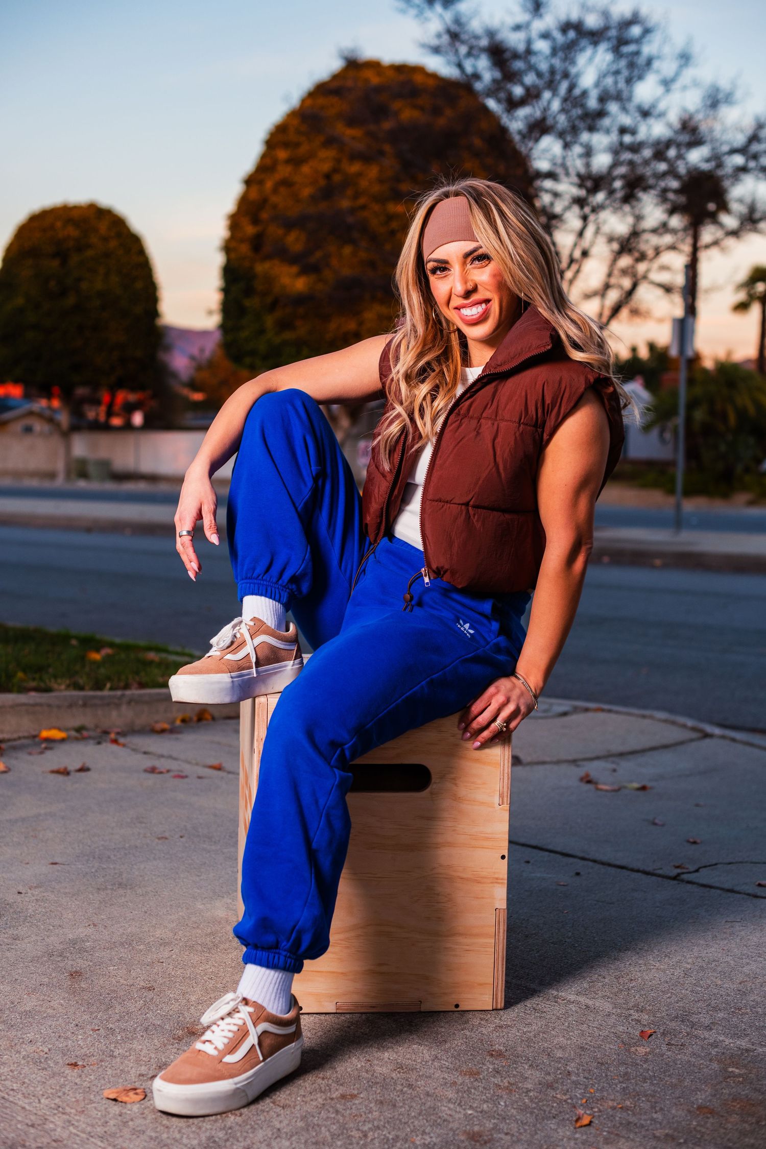 Casual outdoor portrait on wooden box wearing blue pants and brown vest at sunset.