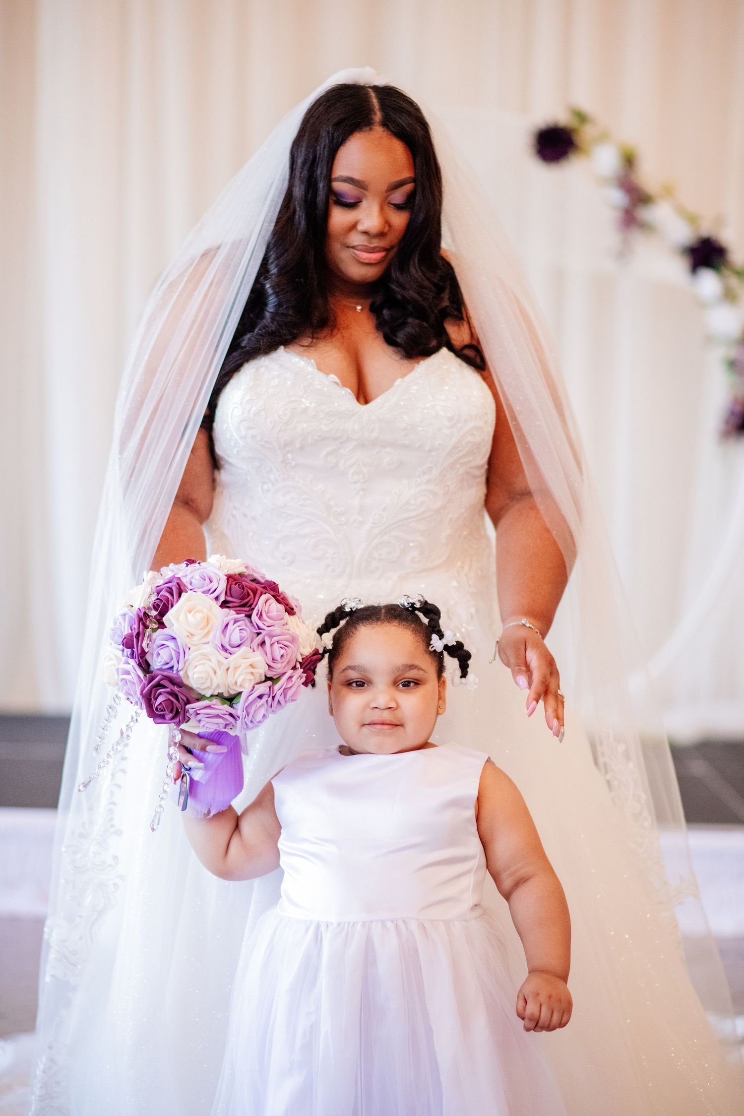 Bride in white dress with young flower girl in matching white dress holding purple bouquet.