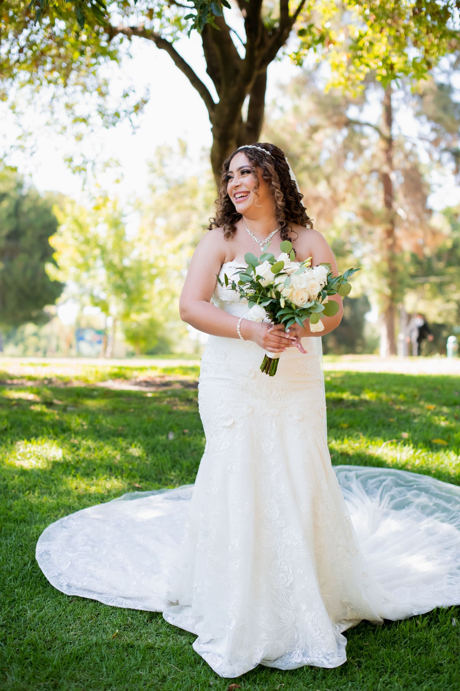 Bride in fitted lace gown holds bouquet while standing on grass under trees.
