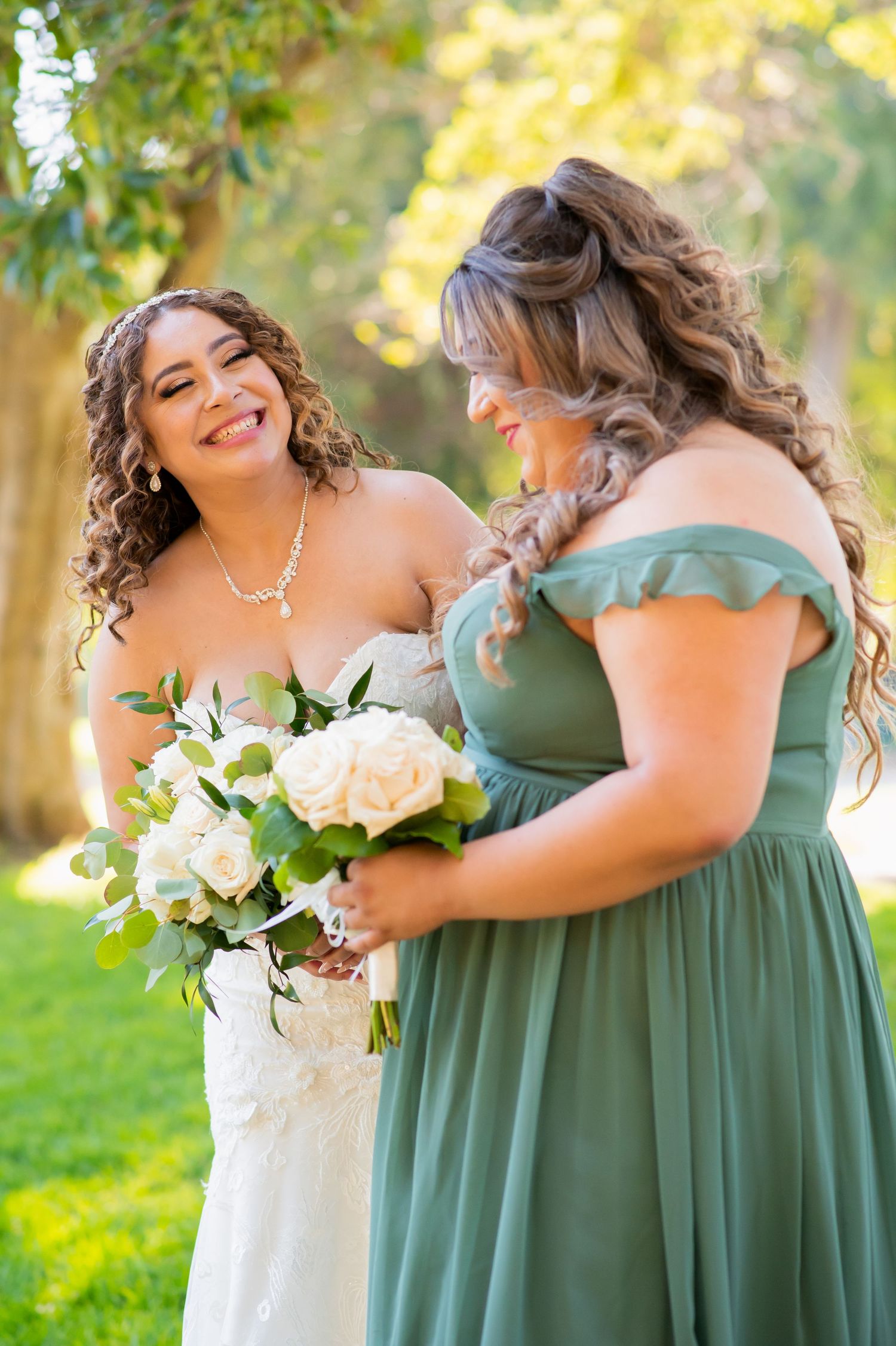 Two wedding party members share a candid moment while holding white rose bouquets in natural lighting.